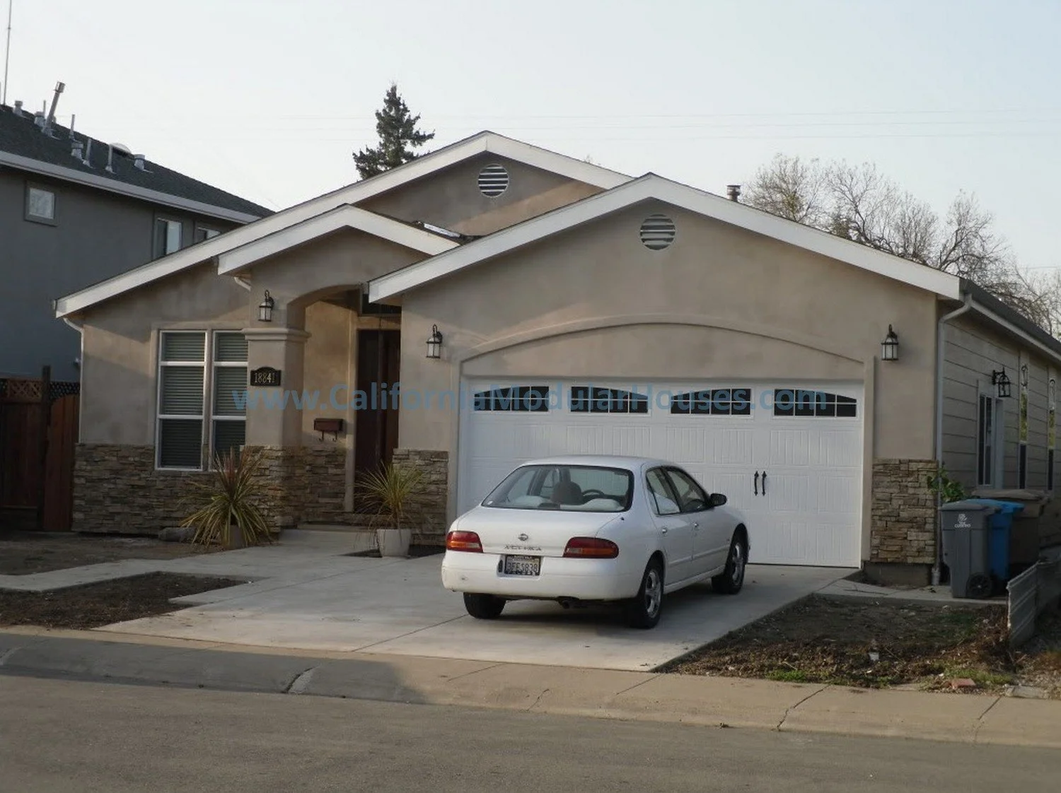 A beige prefab modular house with stone accents, a white garage door, and a small front porch with plants. A white sedan is parked in the driveway.