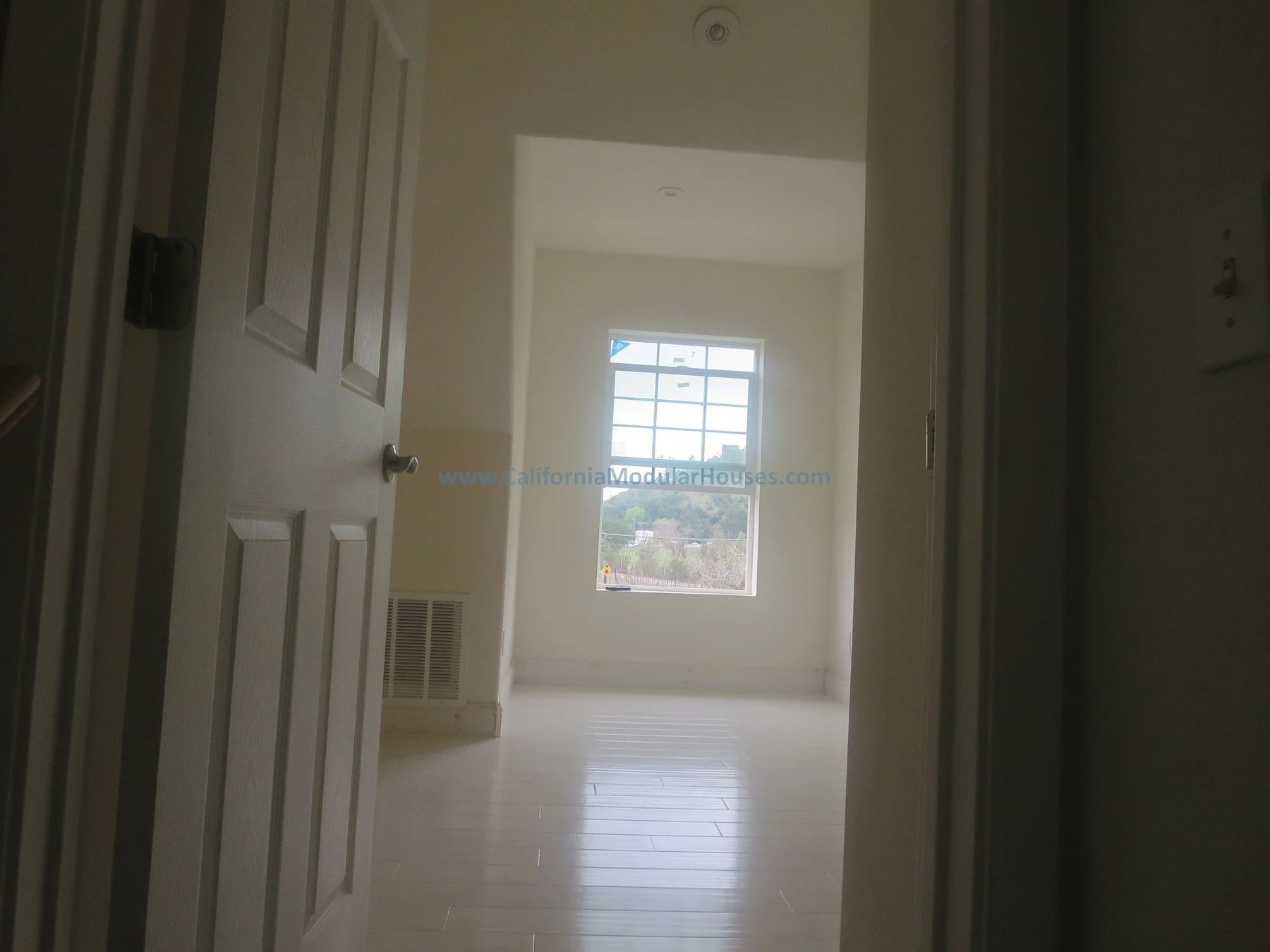 View through an open door into a brightly lit room with a large window, white walls, and light-colored flooring.  Modulars for Napa.  Napa County, CA. Modular. Pre-fab Modular.   Modulars for Napa.  Cape Cod.  