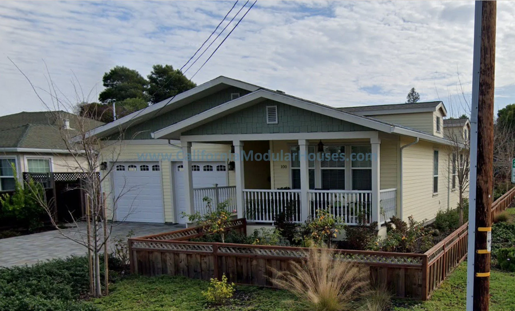 A yellow two-story house with a front porch, white railings, and a brown fenced yard, situated in a suburban neighborhood with trees and cloudy sky.  California Modular Houses,  San Mateo Modular Home,