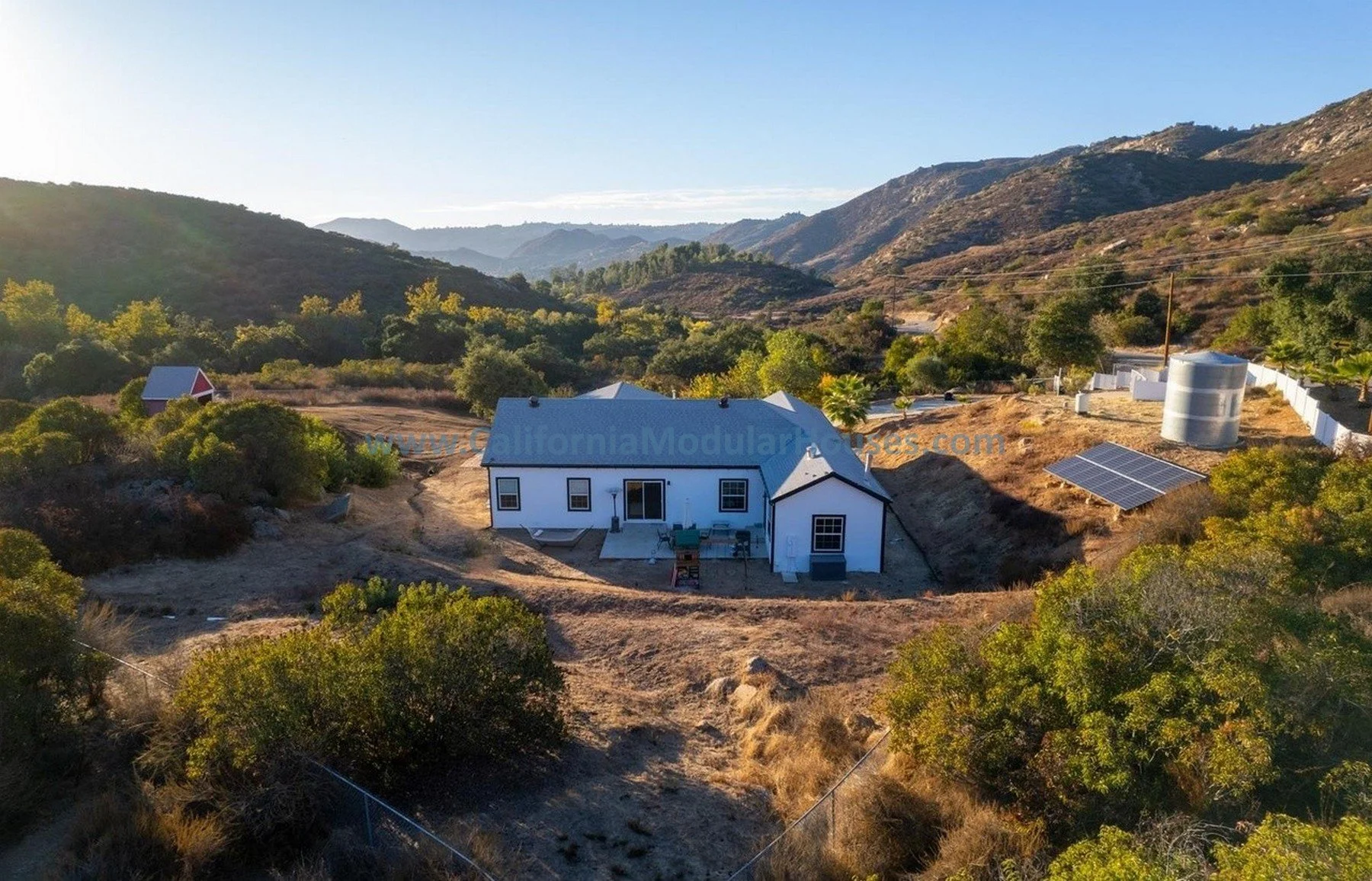 A white house with a gray roof in a hilly, rural landscape surrounded by trees, with solar panels and water tanks nearby and mountains in the background.