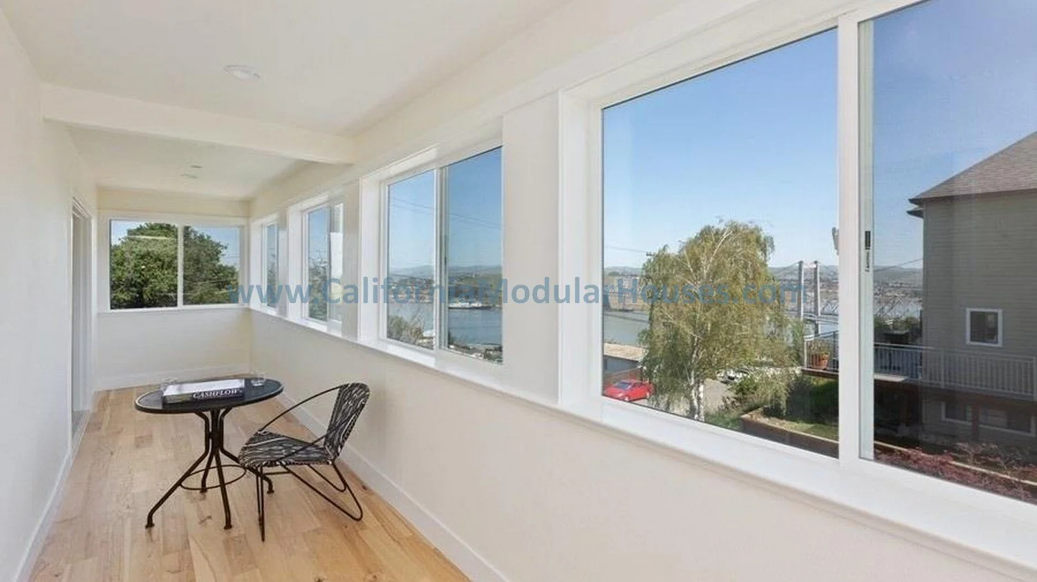 Sunroom with large windows overlooking a river, backyard with trees and neighboring houses, small black table with books, and a black chair.  Contra Costa County 2 story modular home.  Modular house. 