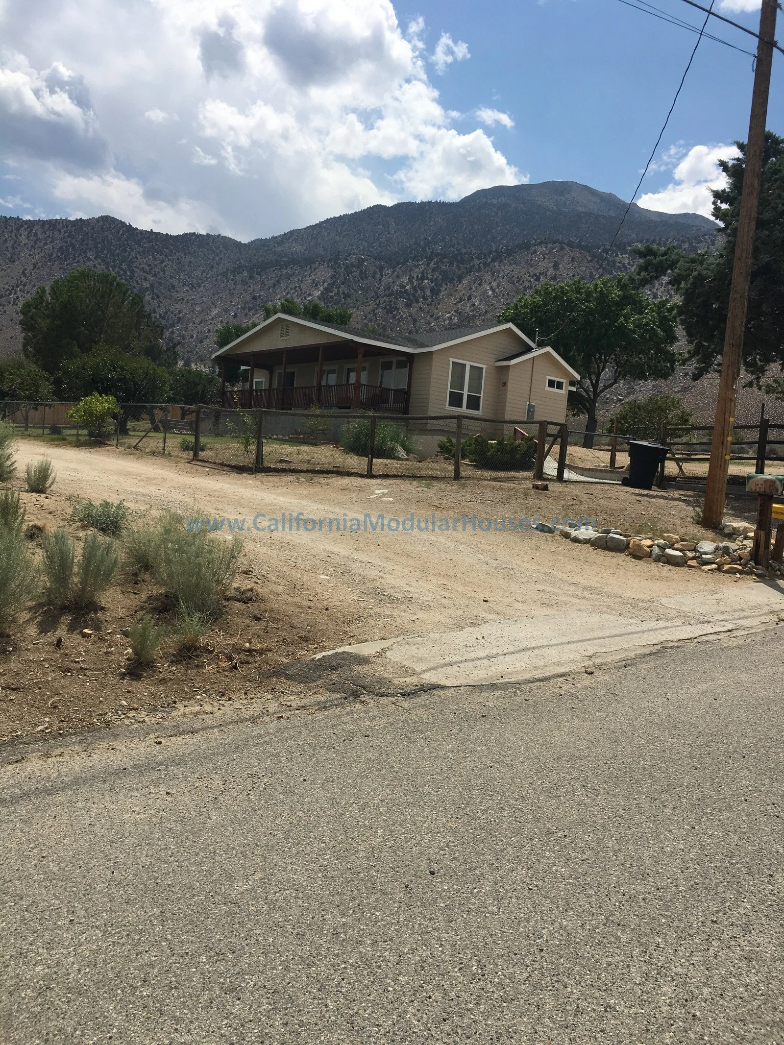 A small house with a porch, surrounded by a wooden fence, situated in a rural area with mountains in the background and a dirt driveway in the foreground.