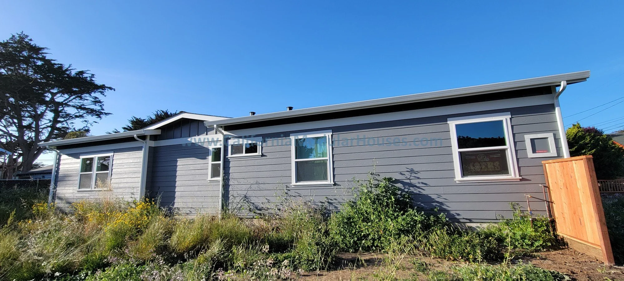 A gray single-story house with white trim, four windows, a small garden with overgrown grass and plants in front, and a partly visible wooden fence on the right side under a clear blue sky.