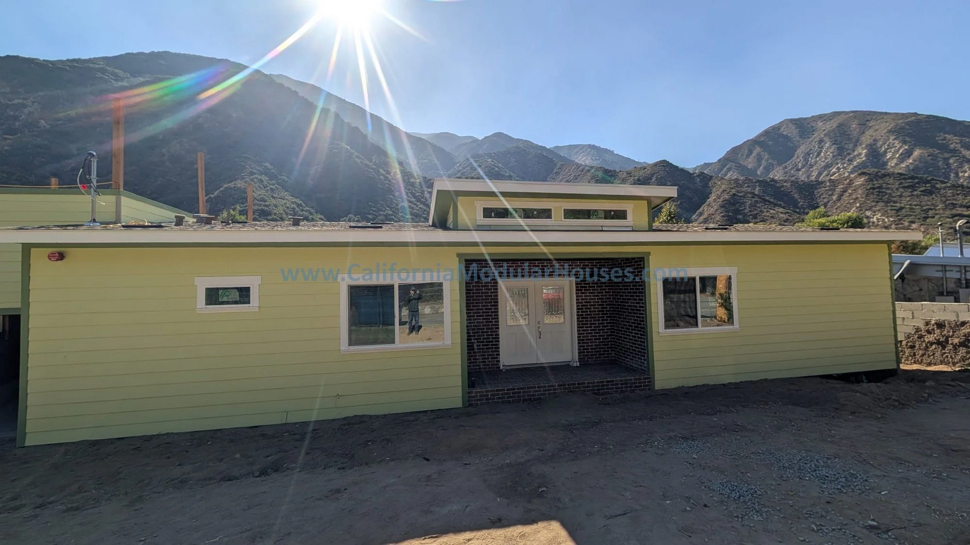 Modern yellow house with white window frames, brick accents around the front door, and a mountain range with the sun shining brightly in the sky behind.