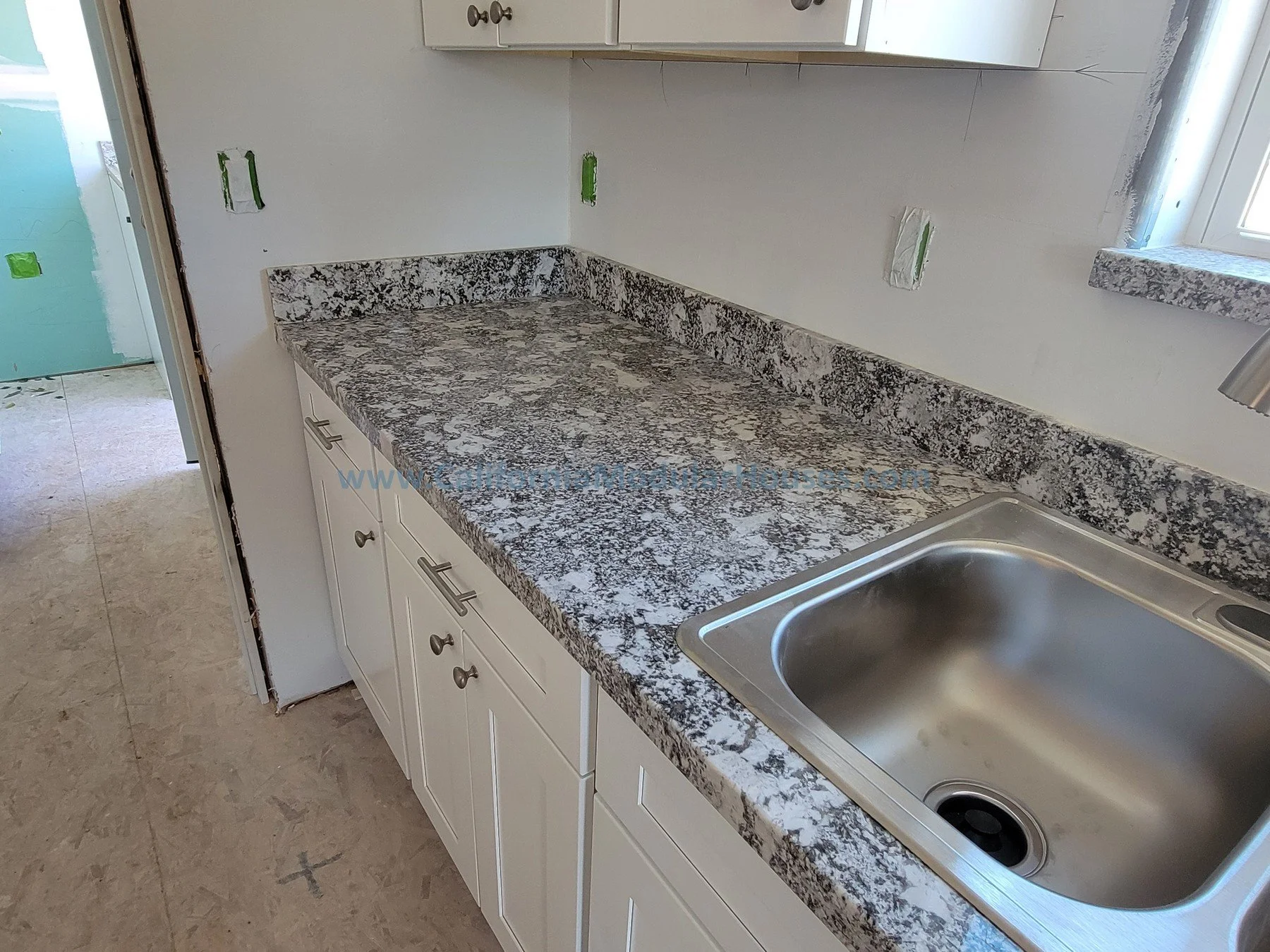 Kitchen countertop with granite surface, white cabinets, stainless steel sink, and electrical outlets on the wall, under a window.