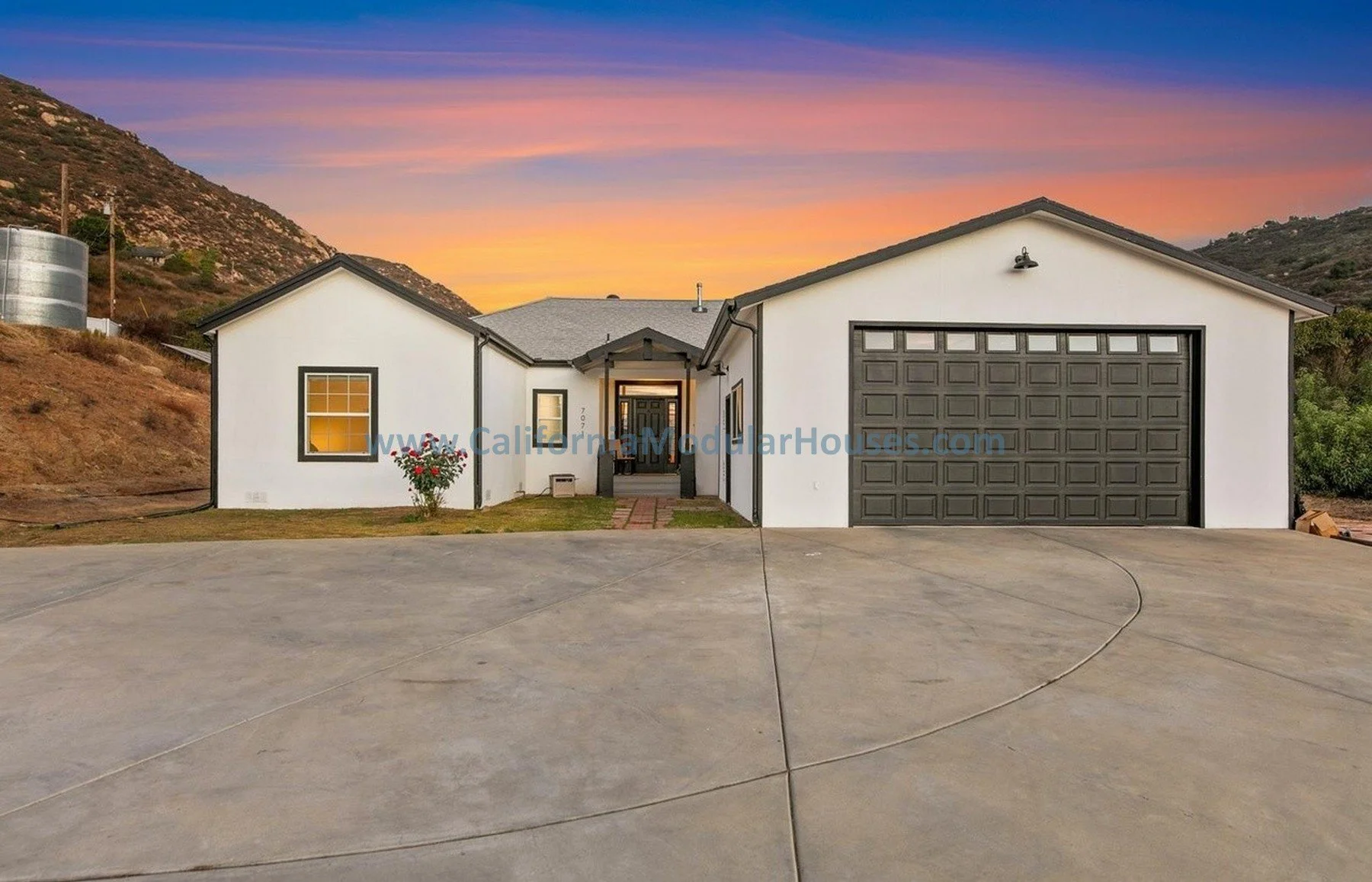 A newly built, white, single-family house with black trim and a dark gray garage door, situated on a concrete driveway at sunset with a colorful sky and hills in the background.