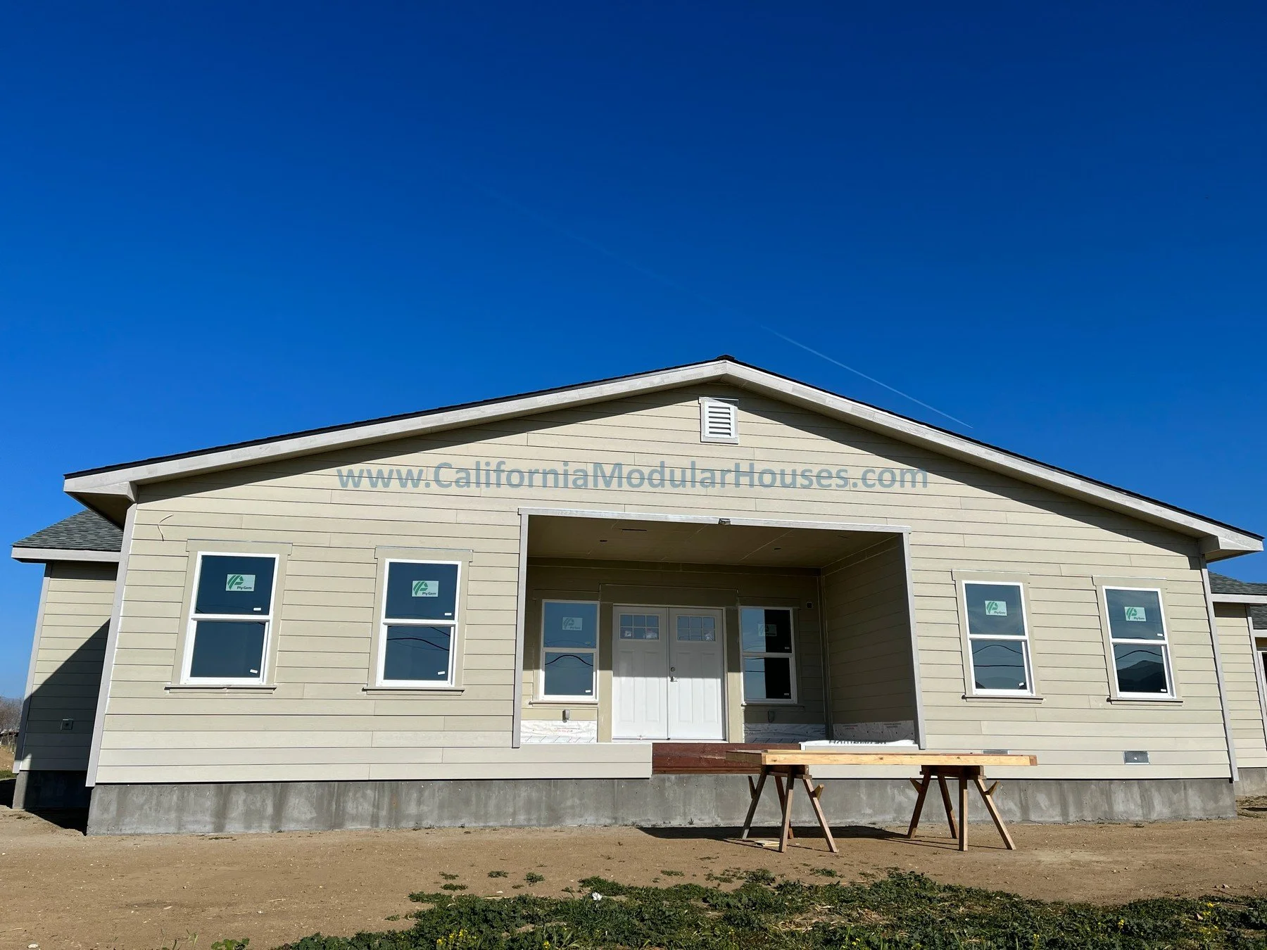 A prefab modular house under construction with beige siding, four windows with stickers, a front door, and a porch area with wooden supports and tools, set against a clear blue sky.  Modular home California