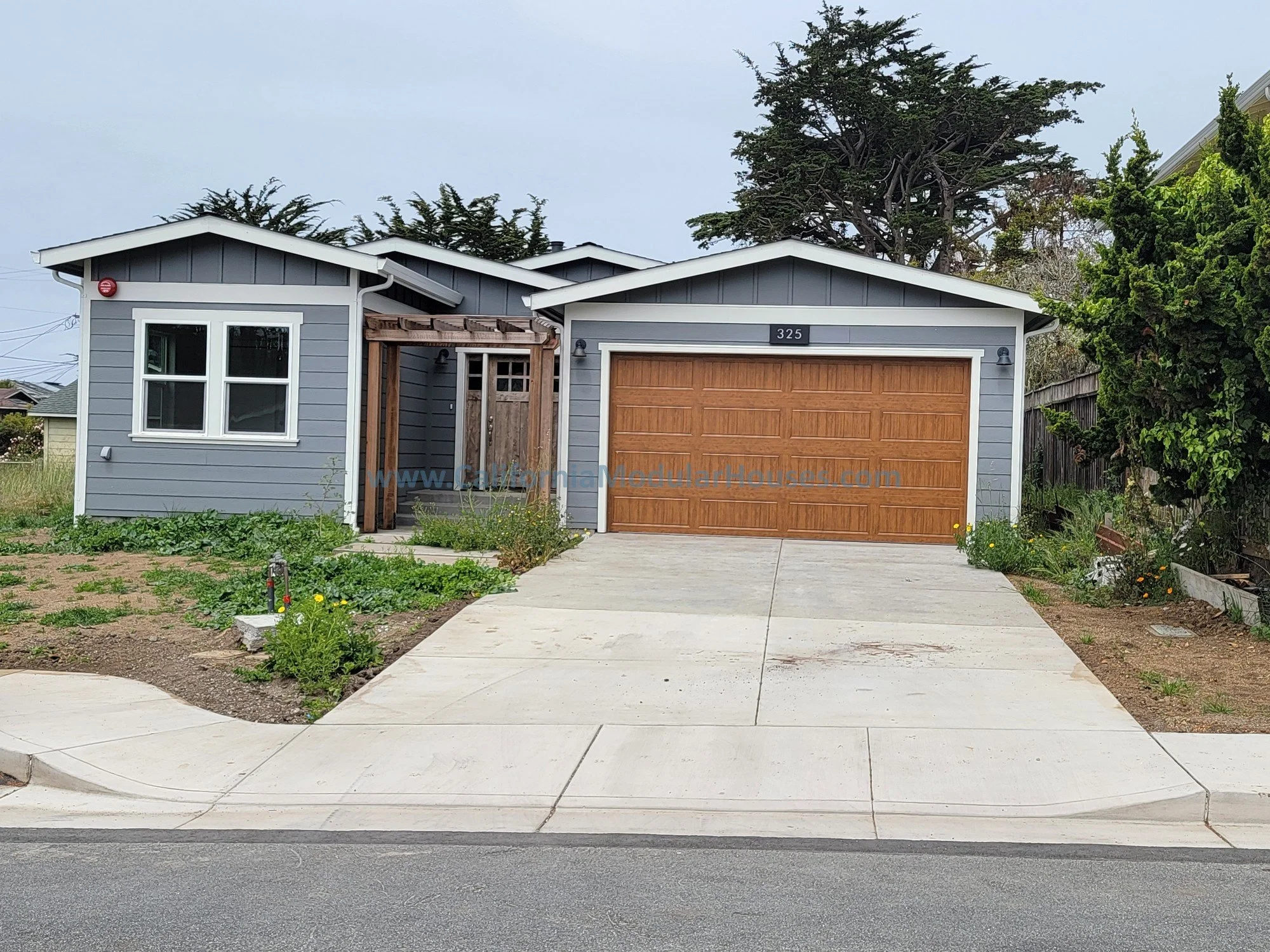 Front view of a modern, single-story house with gray siding, white trim, a wooden garage door, and a concrete driveway. There is a small front porch with a partially constructed wooden pergola and some plants along the walkway.