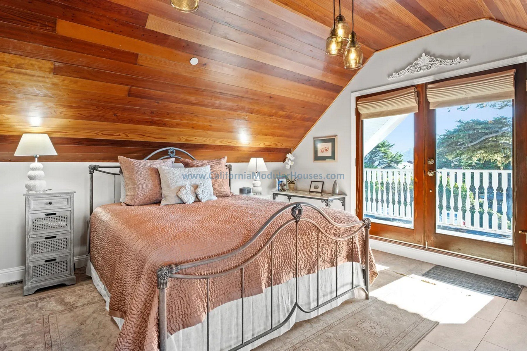 A cozy bedroom with a metal bed frame, plush pink bedding.  large window with wooden framing and a balcony outside, wooden ceiling with hanging lights, and a beige tiled floor with a rug.  Modular residence.  Prefab CA