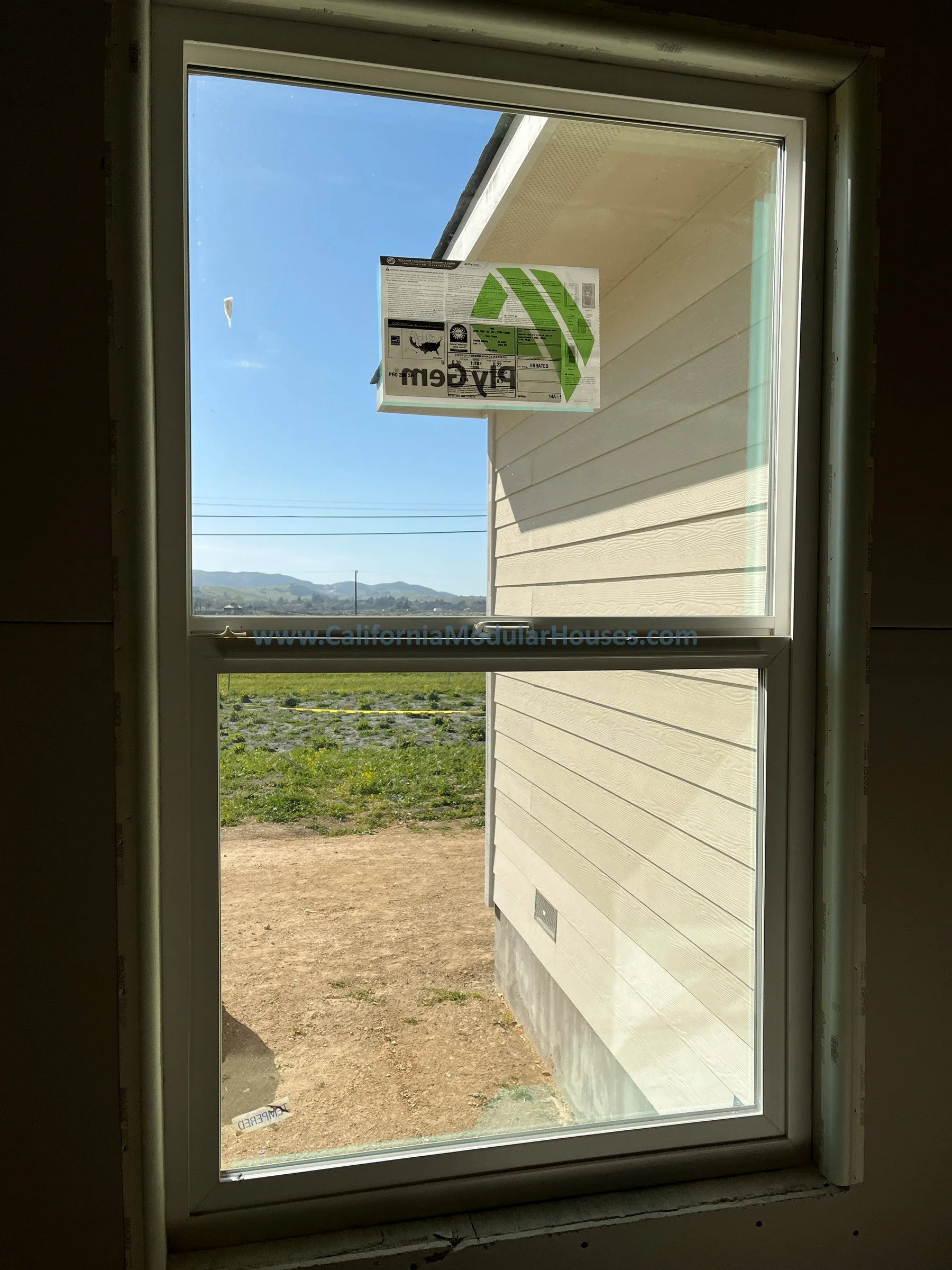 View of the outdoors through a new double-pane window, showing a house siding, a grassy yard, and distant hills with a clear blue sky.  Modular CA.  California modular home.  