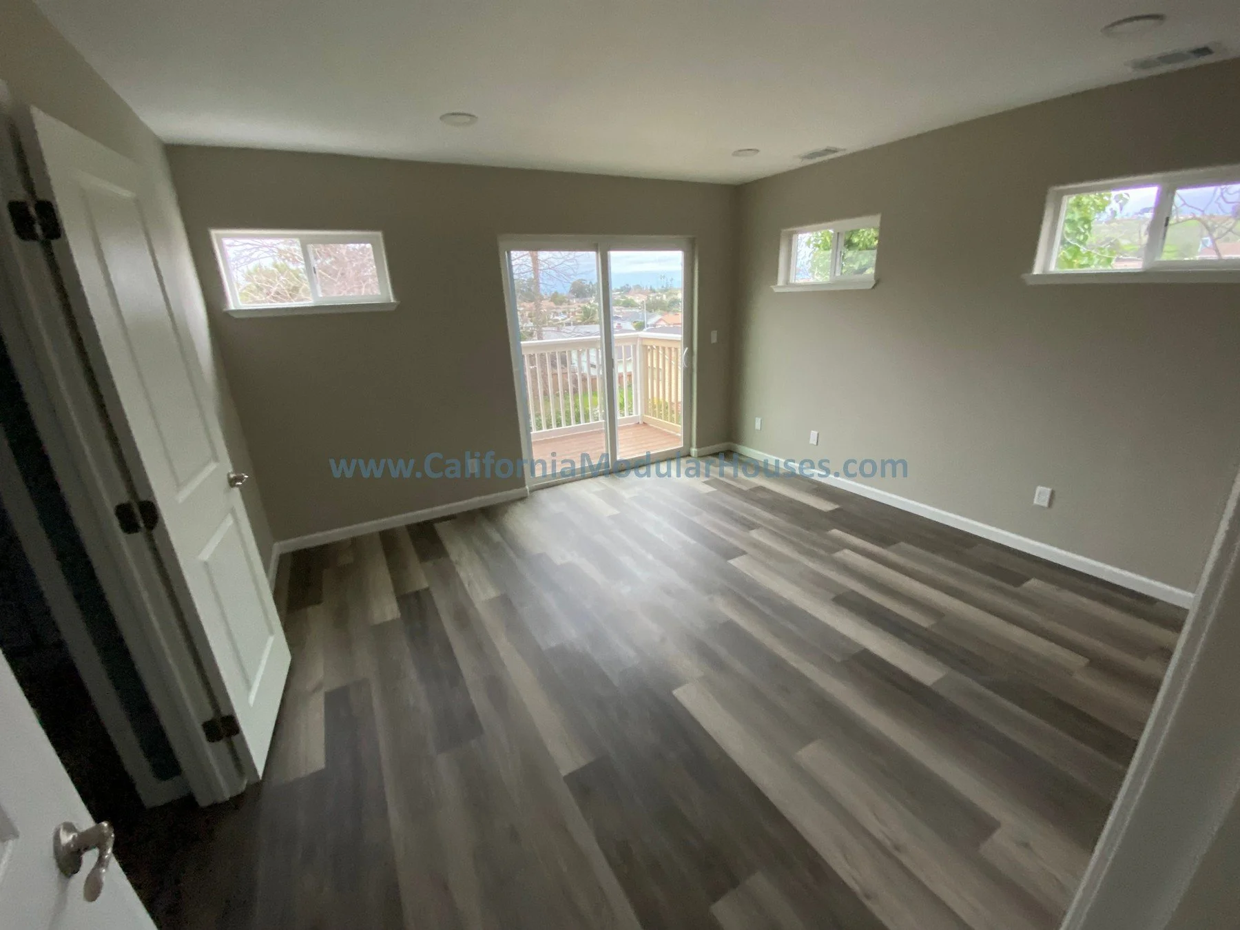 Empty bedroom with light grey walls, dark and light patterned wood flooring, four rectangular windows, a glass sliding door leading to a balcony, and a ceiling with recessed lighting.  Prefab modular ADU