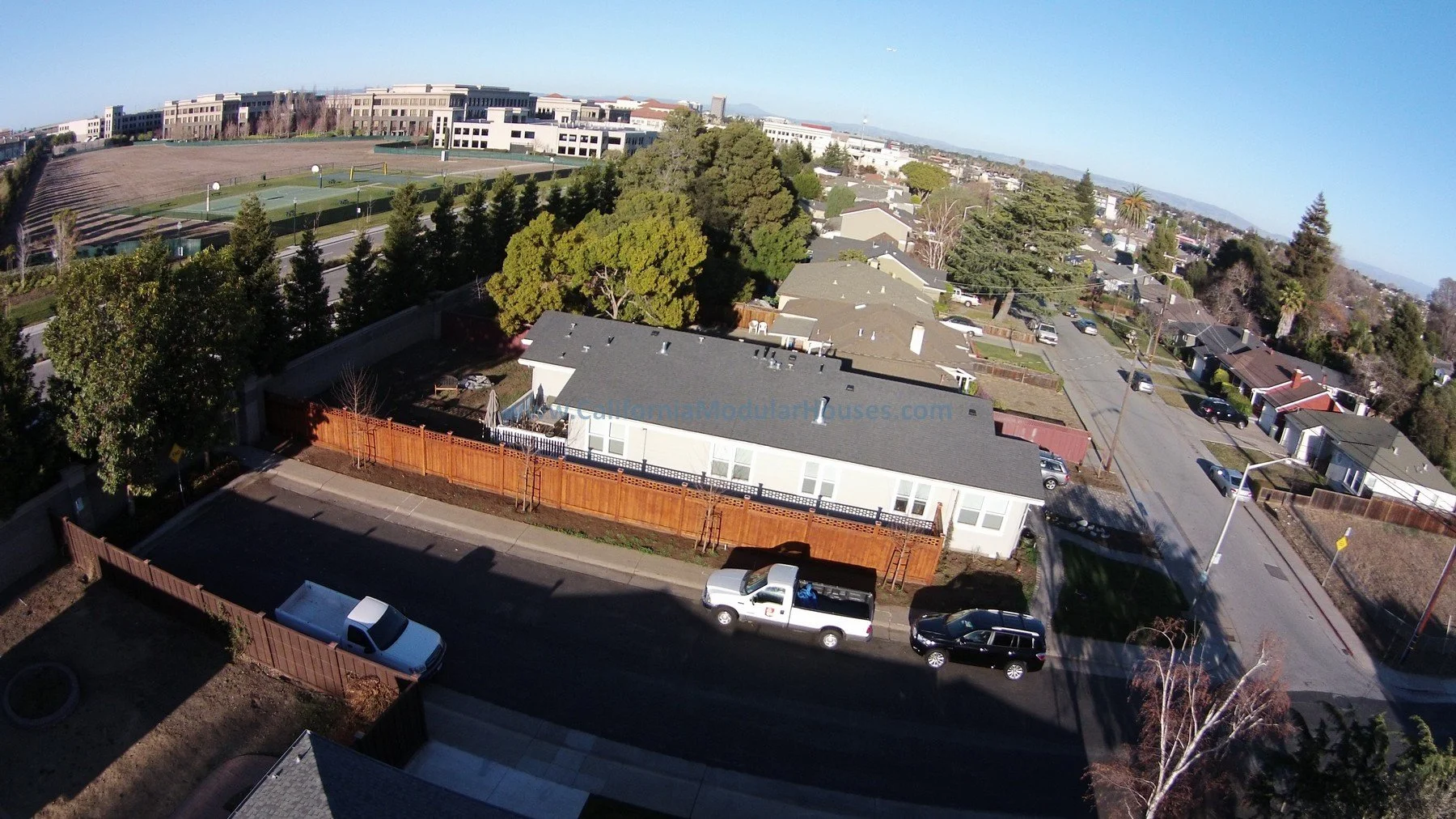 Aerial view of a residential neighborhood with various houses, trees, parked cars on the street, and a sports complex with tennis courts in the background under a clear sky.  San Francisco Bay Area, CA.  Prefab Houses, Prefab Homes.