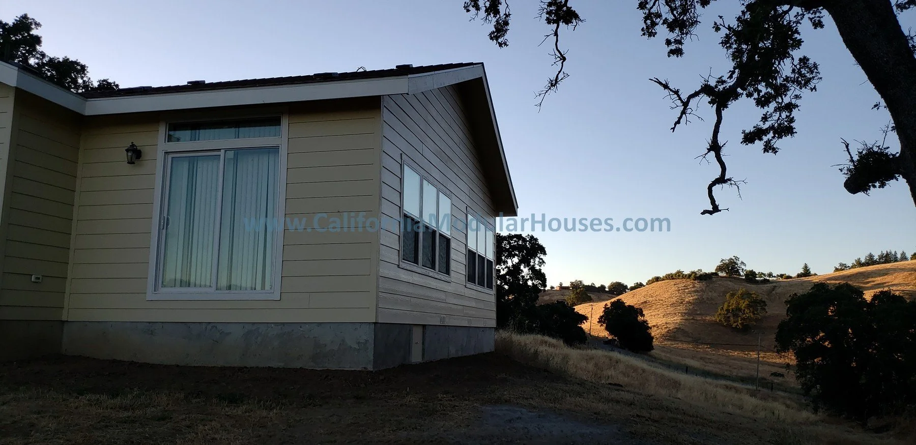 Side view of a beige house with large windows, situated on a hilly landscape during sunset with trees and hills in the background. California Modular Houses, Modular Home on a Sloped Property. 