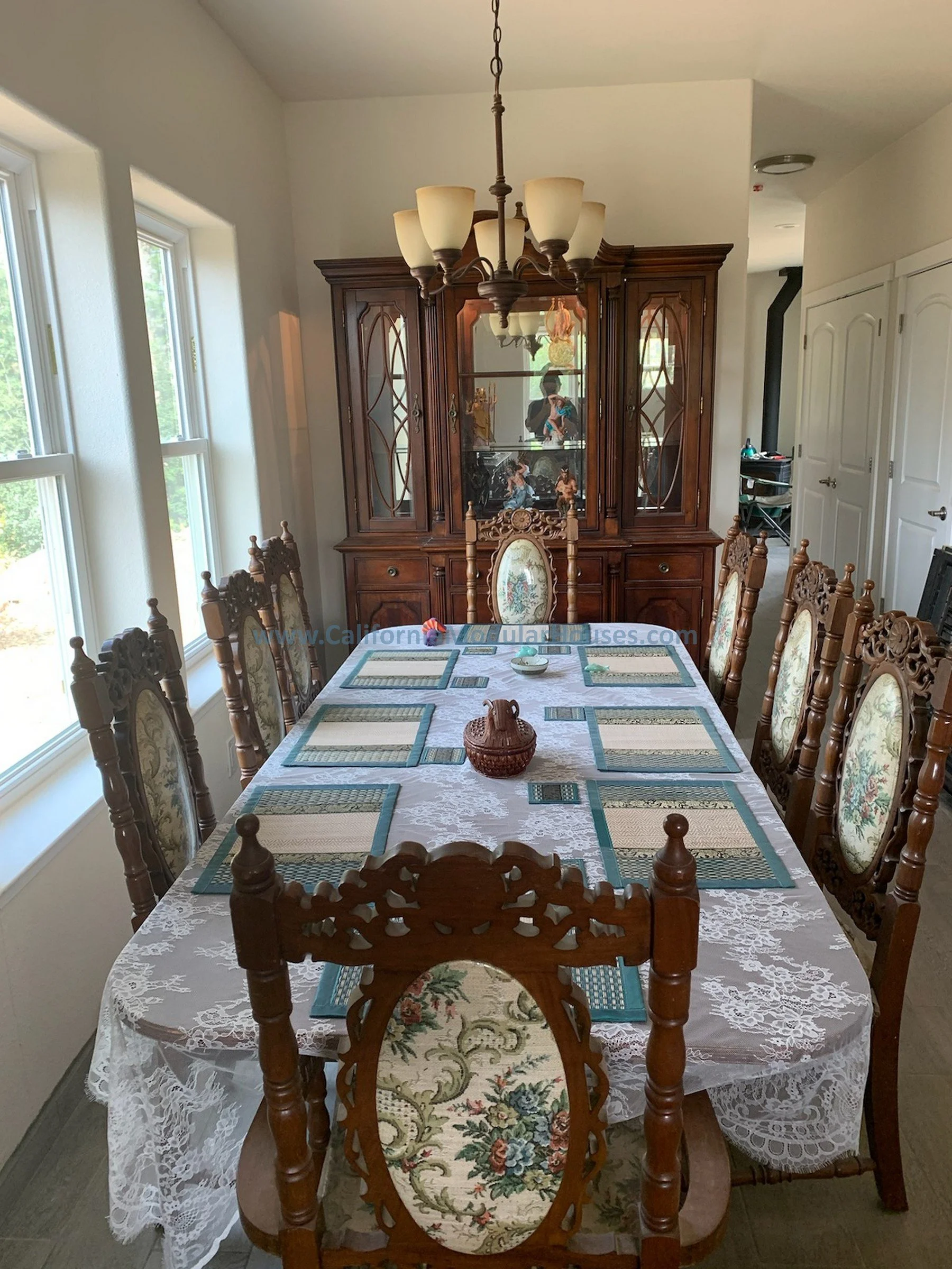 A dining room with a large wooden table covered with a lace tablecloth, surrounded by eight ornate wooden chairs with floral upholstery.  The room has three windows on the left, providing natural light.  California Modular, Modular Home CA,