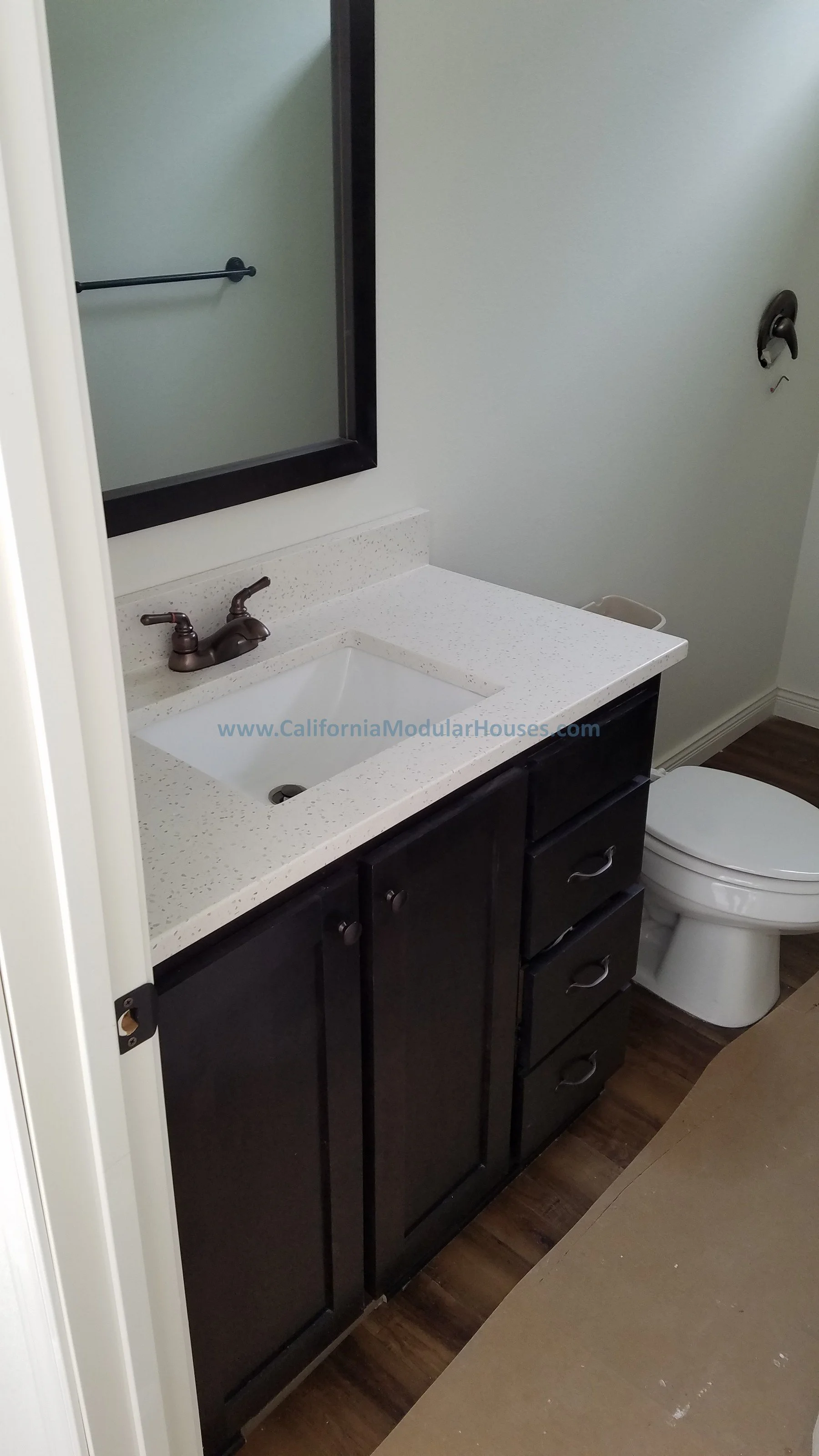 Bathroom vanity with a black cabinet, a white speckled countertop, a built-in sink, and a bronze faucet. A mirror is mounted above the sink, and a toilet is visible next to the vanity. City of Santa Barbara, Santa Barbara County, CA.  Modular.
