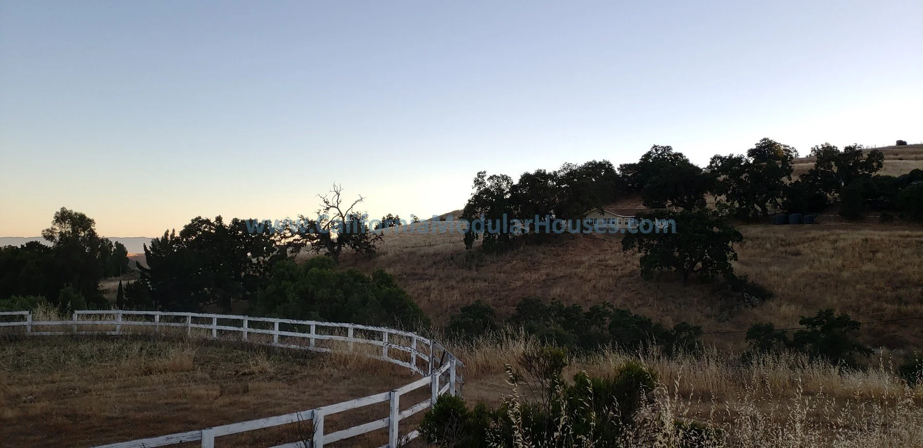 Hilly landscape with dry grass and scattered trees, white wooden fence in the foreground, and a house on the hill. The sky is clear, likely near sunset or sunrise. San Jose Modular Home, Modular Home CA,