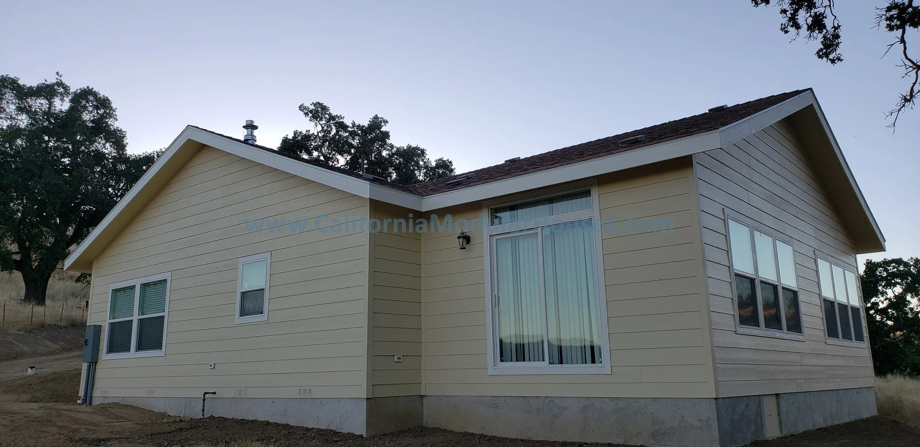 A beige house with a sloped roof and multiple windows, set in a rural area with trees and open fields in the background.  Pre-Fabricated Homes, Prefab Home California, Modular Homes in California.