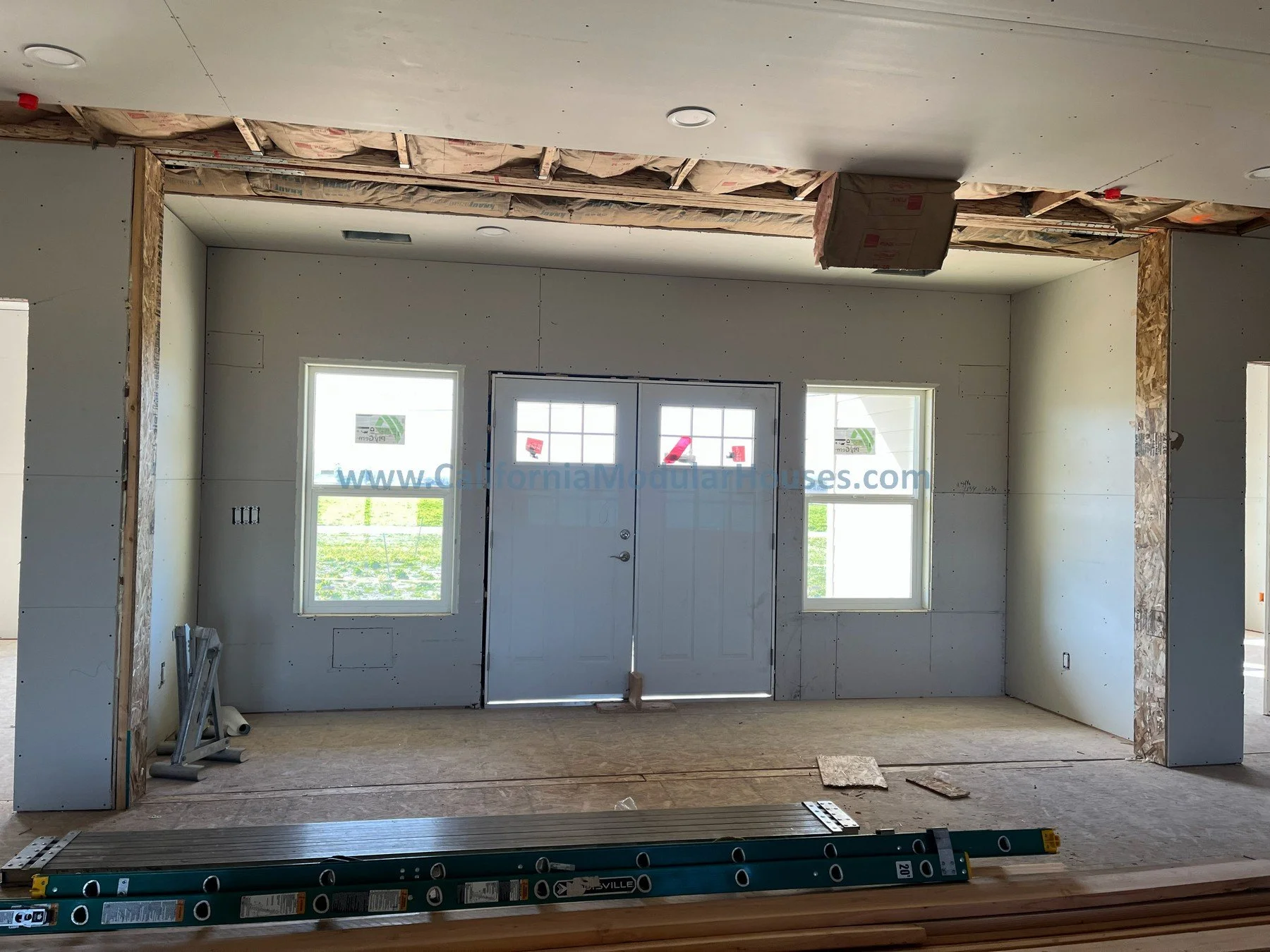 Interior view of a house under construction, featuring a doorway with a door and two windows, with visible drywall and construction materials.