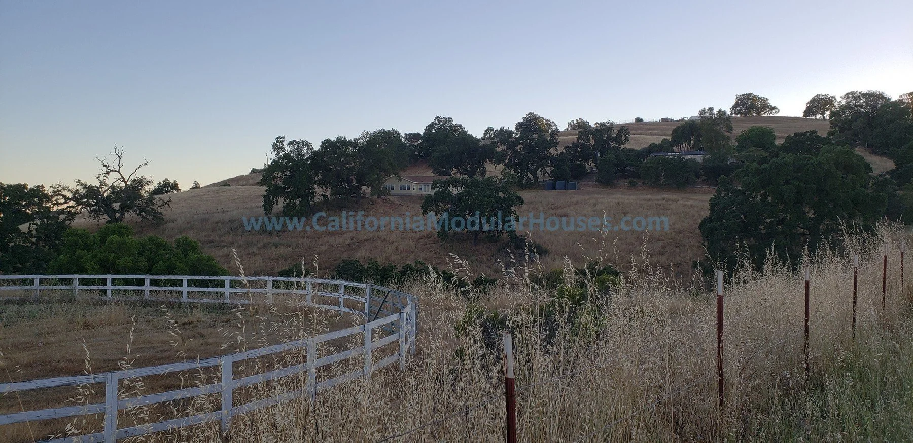A rural landscape with rolling hills, trees, and dry grasses. There is a white fence in the foreground and a house on top of the hill in the distance. The sky is clear and blue.  Bay Area Prefab Modular Home, California Modular Homes,