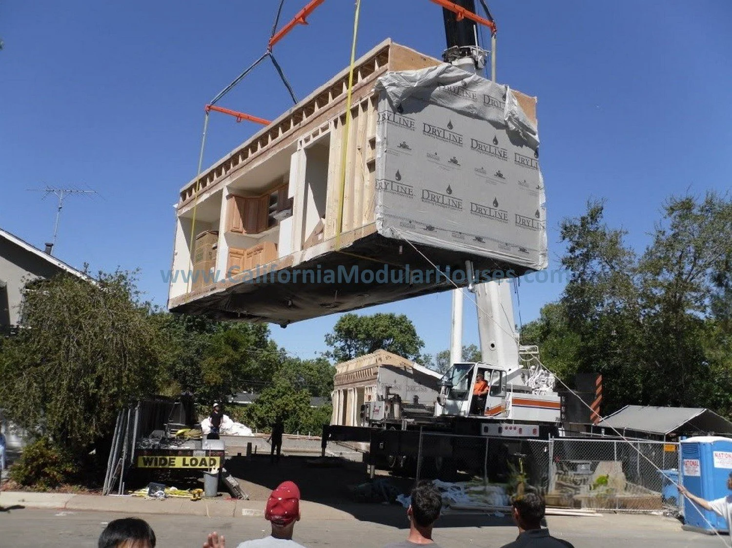 A construction site with a large crane lifting a modular home. The modular home is being installed on a foundation, with workers on the ground observing. The sky is clear and blue, and there are trees and other buildings nearby.