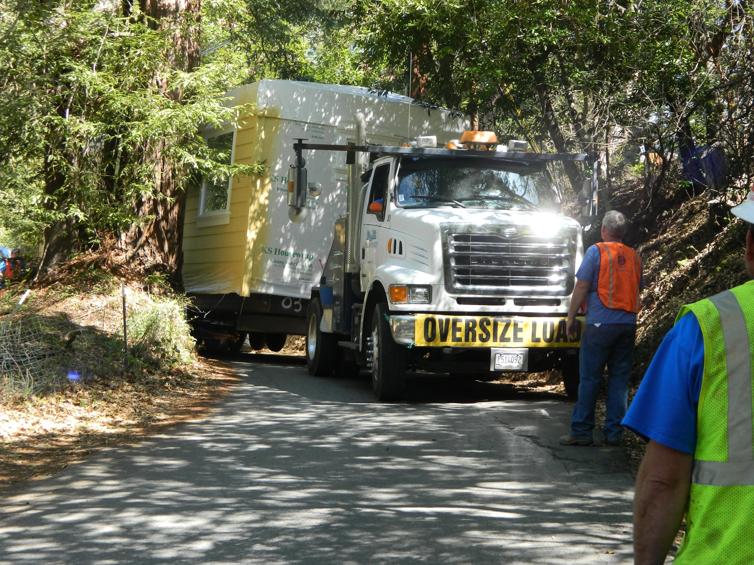 Large truck with 'OVERSIZE LOAD' on the front is on a narrow, winding forested road, with trees and branches on both sides. Several workers wearing safety vests are inspecting the situation and verifying that the home is delivered safely.