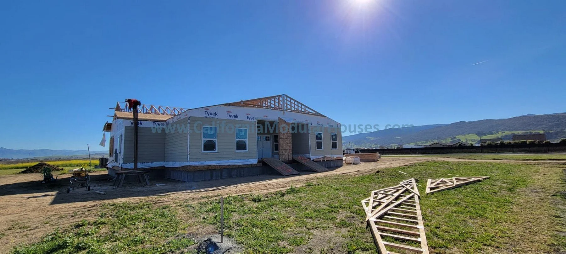 Under construction house on a cleared lot with wooden framing on roof, windows installed, and building materials and tools on site, under clear blue sky with mountains in background.  Prefab modular.  California.  