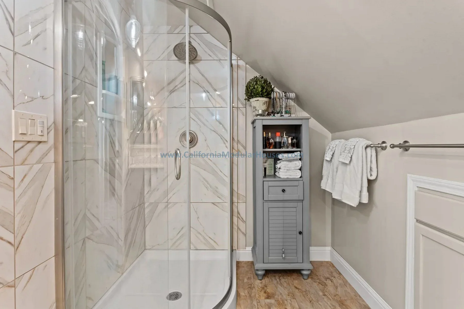 Bathroom with a curved glass shower stall on the left, gray storage cabinet with towels and toiletries in the center, white towels hanging on a towel bar on the right, and a hardwood floor.  Prefab house for Mendocino, California.  