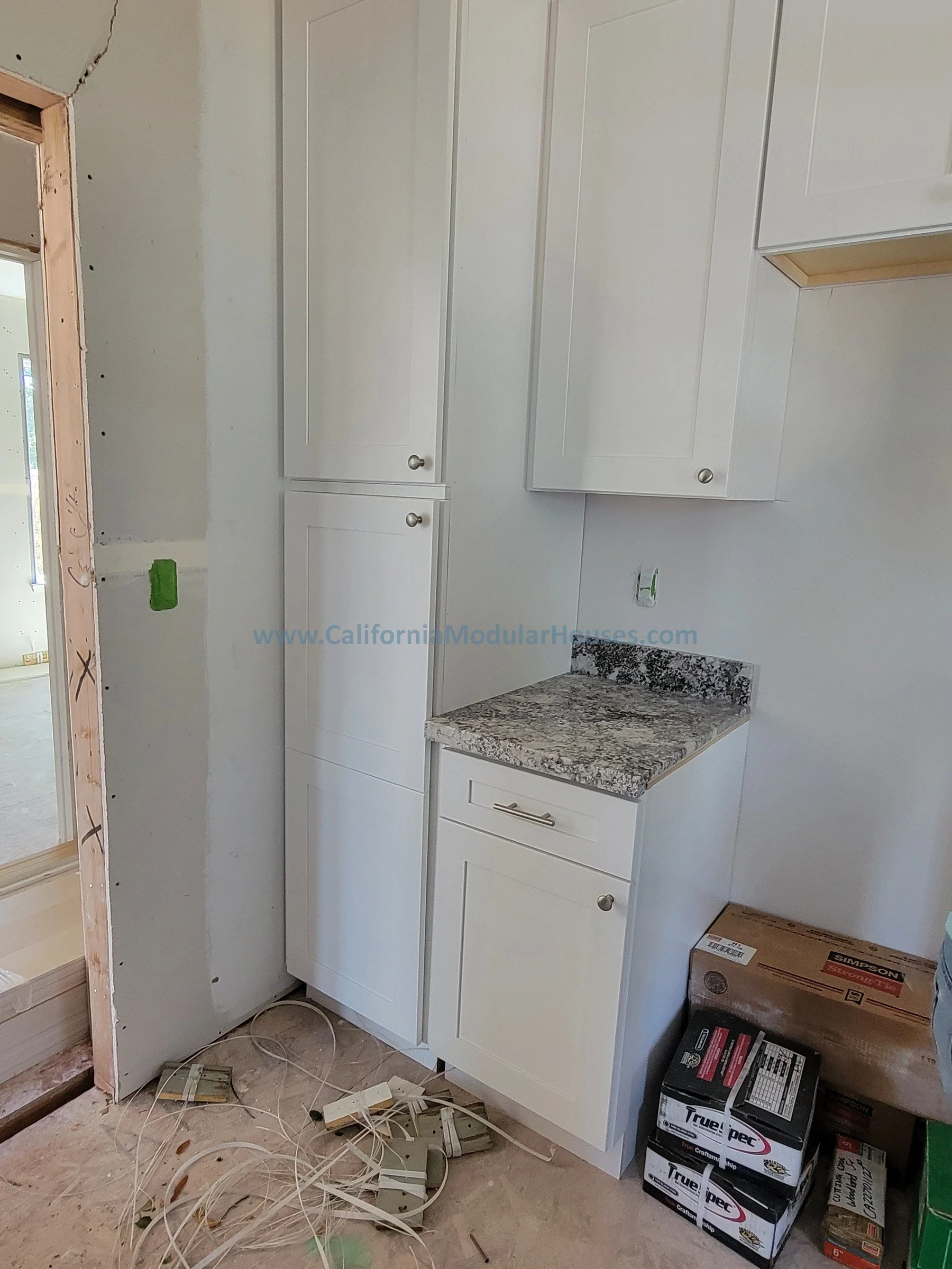 Under construction kitchen corner with white cabinets, granite countertop, and construction materials on the floor.