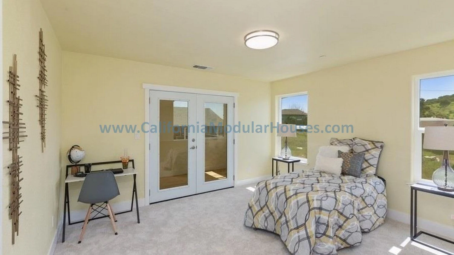 Furnished bedroom with a bed, two side tables with lamps, a small desk with a chair, and a glass door leading outside, with windows showing a green landscape.  California Modular Houses, Inc.  