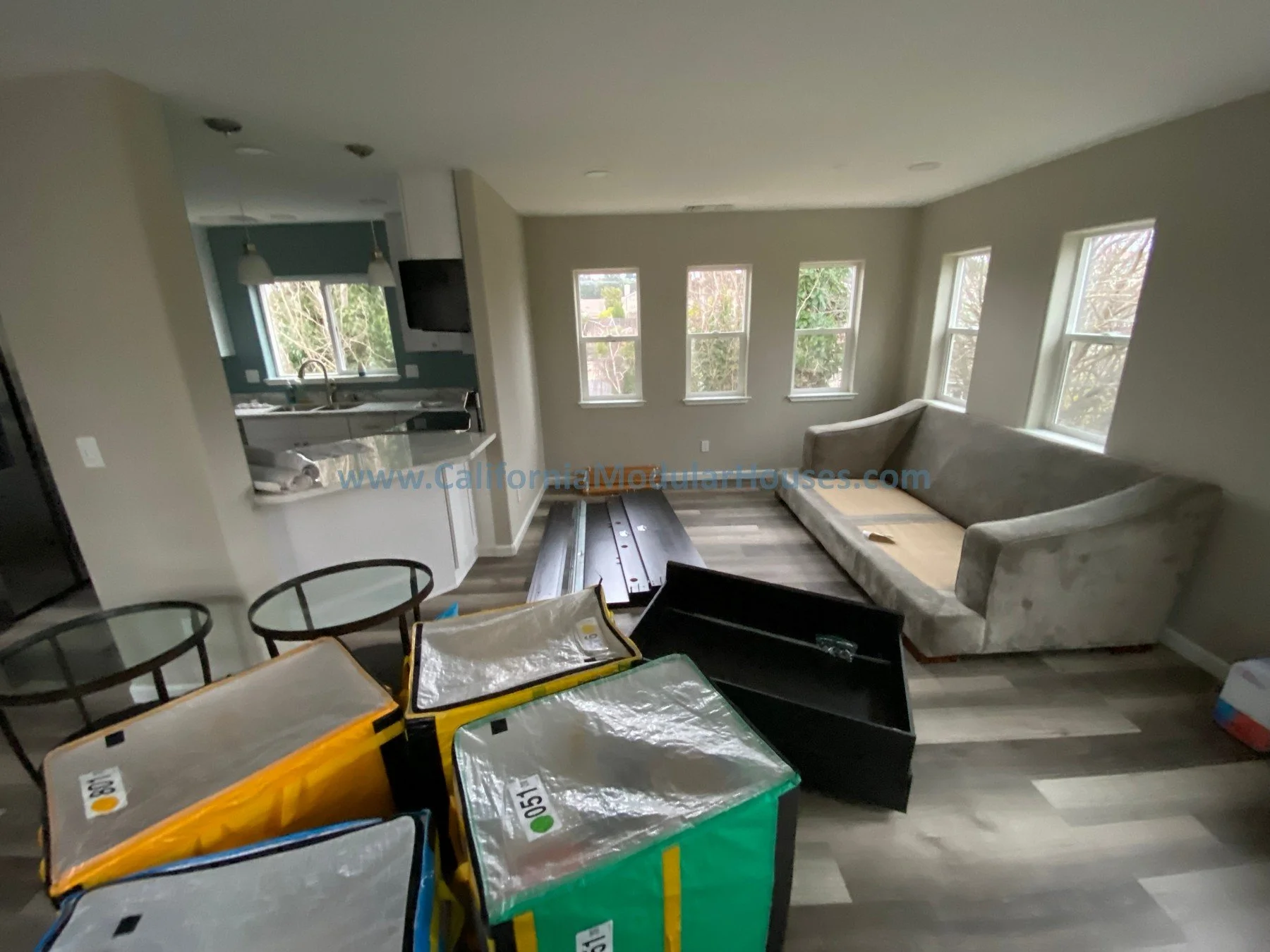 Living room with a gray couch, multiple windows, and packs of flooring materials on the floor, near a dining table and in front of an open kitchen.  Prefab modular.    Modular Contra Costa County, CA.  