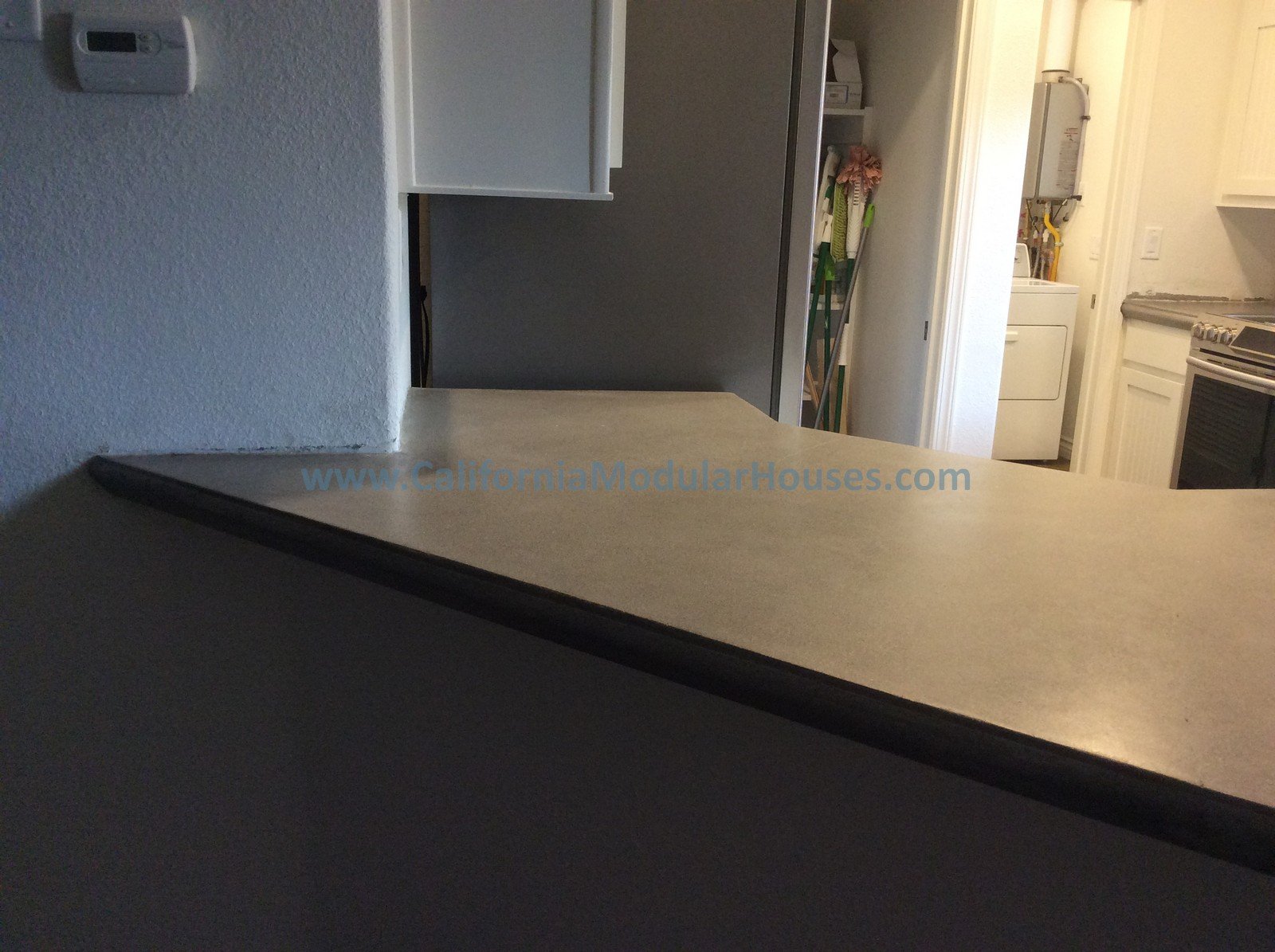 View of a kitchen counter with a website URL printed on it, showing appliances and cleaning tools in the background.