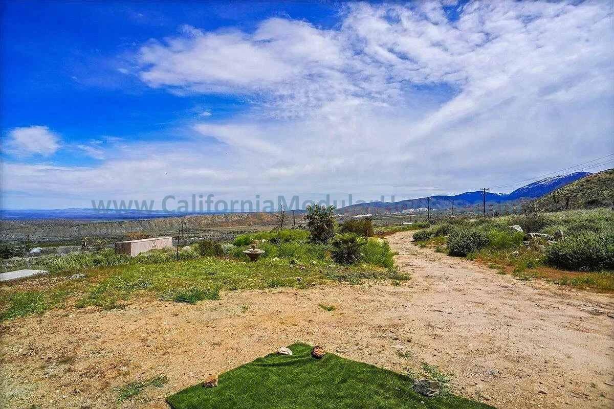 A dirt pathway running through a grassy landscape with a few plants and rocks, extending towards distant hills and mountains under a partly cloudy sky.