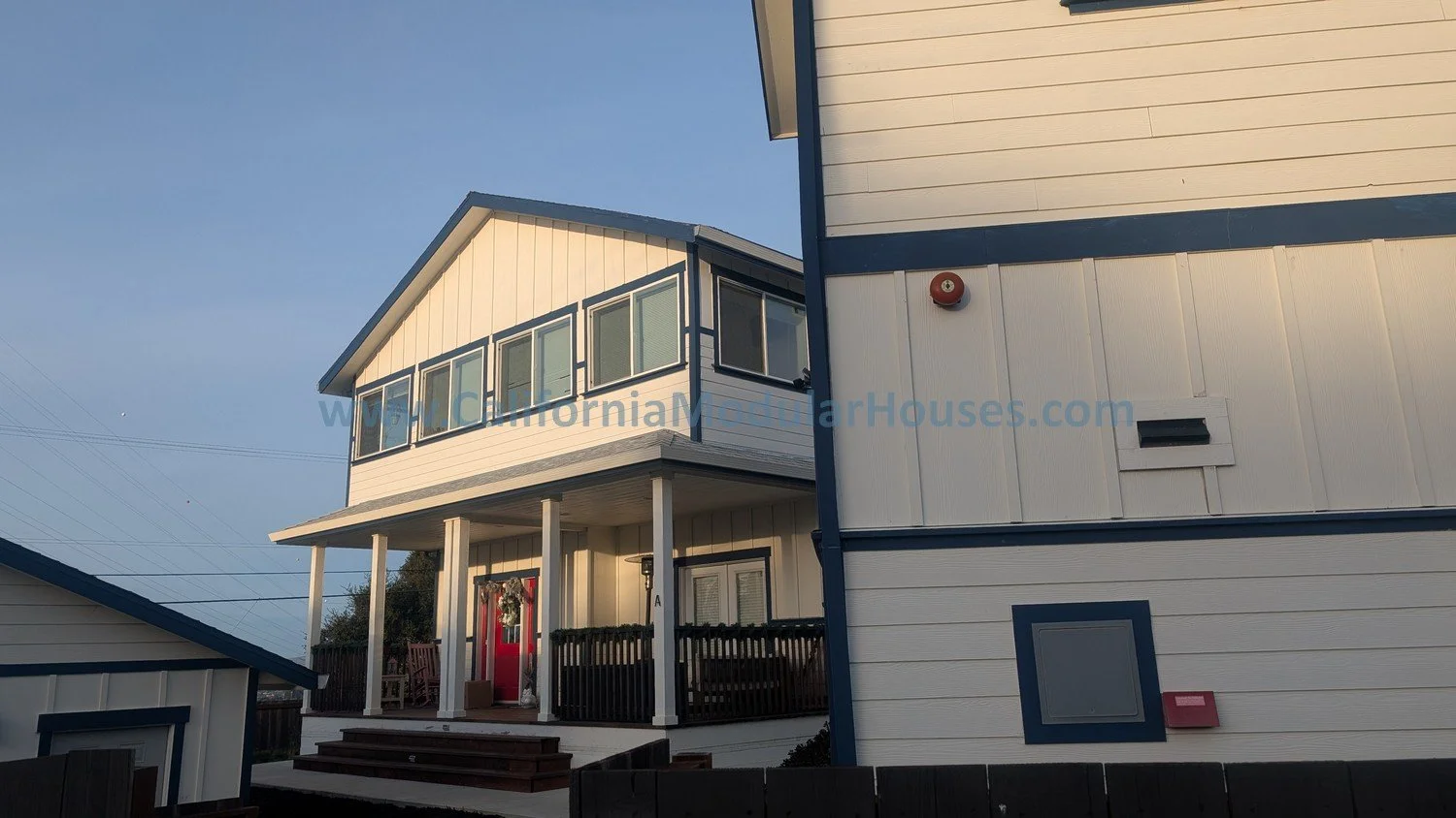 White two-story house with a red front door, porch, and a wreath, with a small staircase leading up to it, under a clear blue sky.