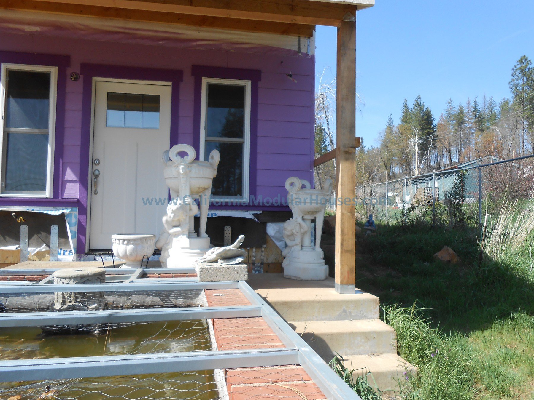 Front porch of a purple house with white door and windows, decorative white urns with cherub figures, concrete steps, and construction materials in the yard, surrounded by a grassy area and trees.
