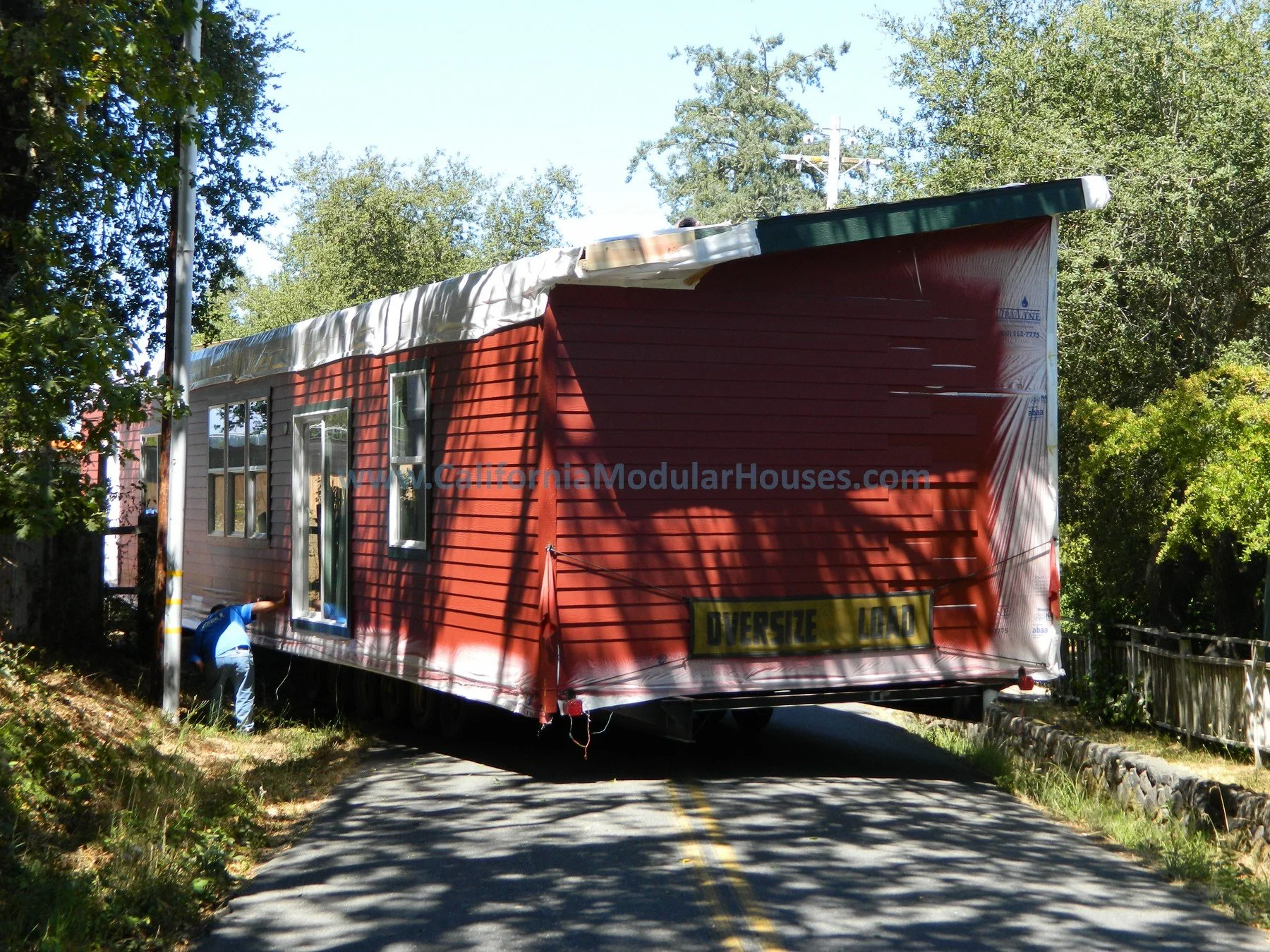 A red modular house being transported on a trailer truck on a narrow two-lane road surrounded by trees. Prefab Home California, Pre-Fabricated Homes.