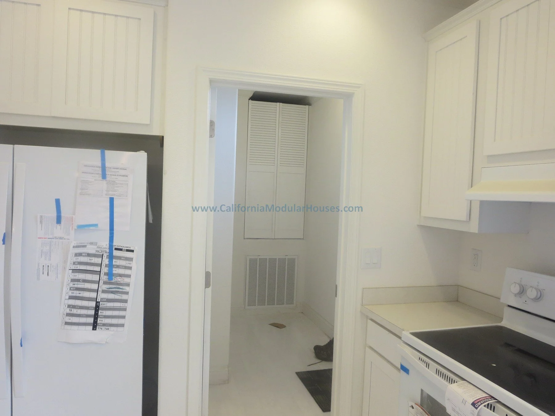 Kitchen with white cabinets, a white stove, and a refrigerator with papers taped on it. The image shows a doorway leading to another room with louvered closet doors. California modular home.   Unfinished attic.  New prefab modular house in Napa, CA. 