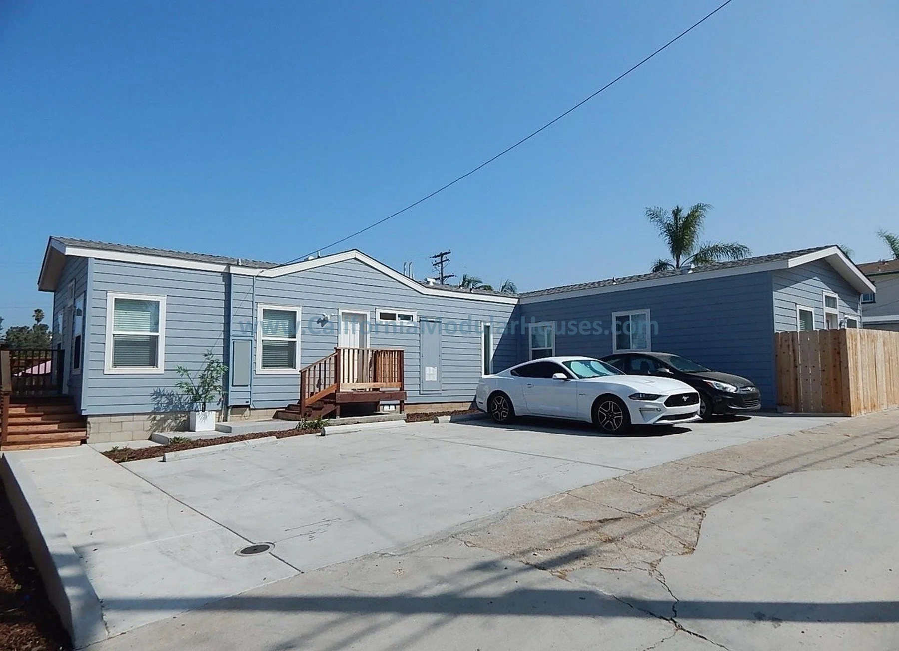 A light blue house with white trim, two cars parked in front, small wooden stairs leading to the front door, and a concrete driveway under a clear blue sky.