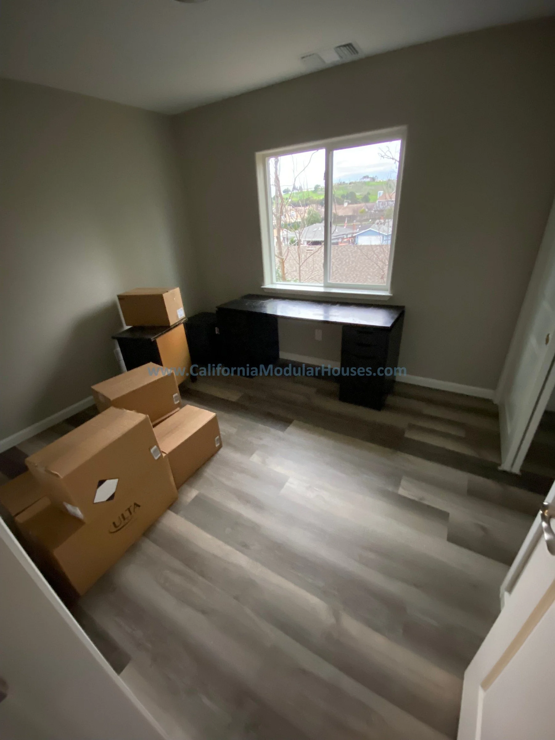 Empty bedroom with light gray walls and wood laminate flooring, containing four cardboard boxes in the corner, a black desk beneath a window.  California modular.   Contra Costa County California Modular ADU