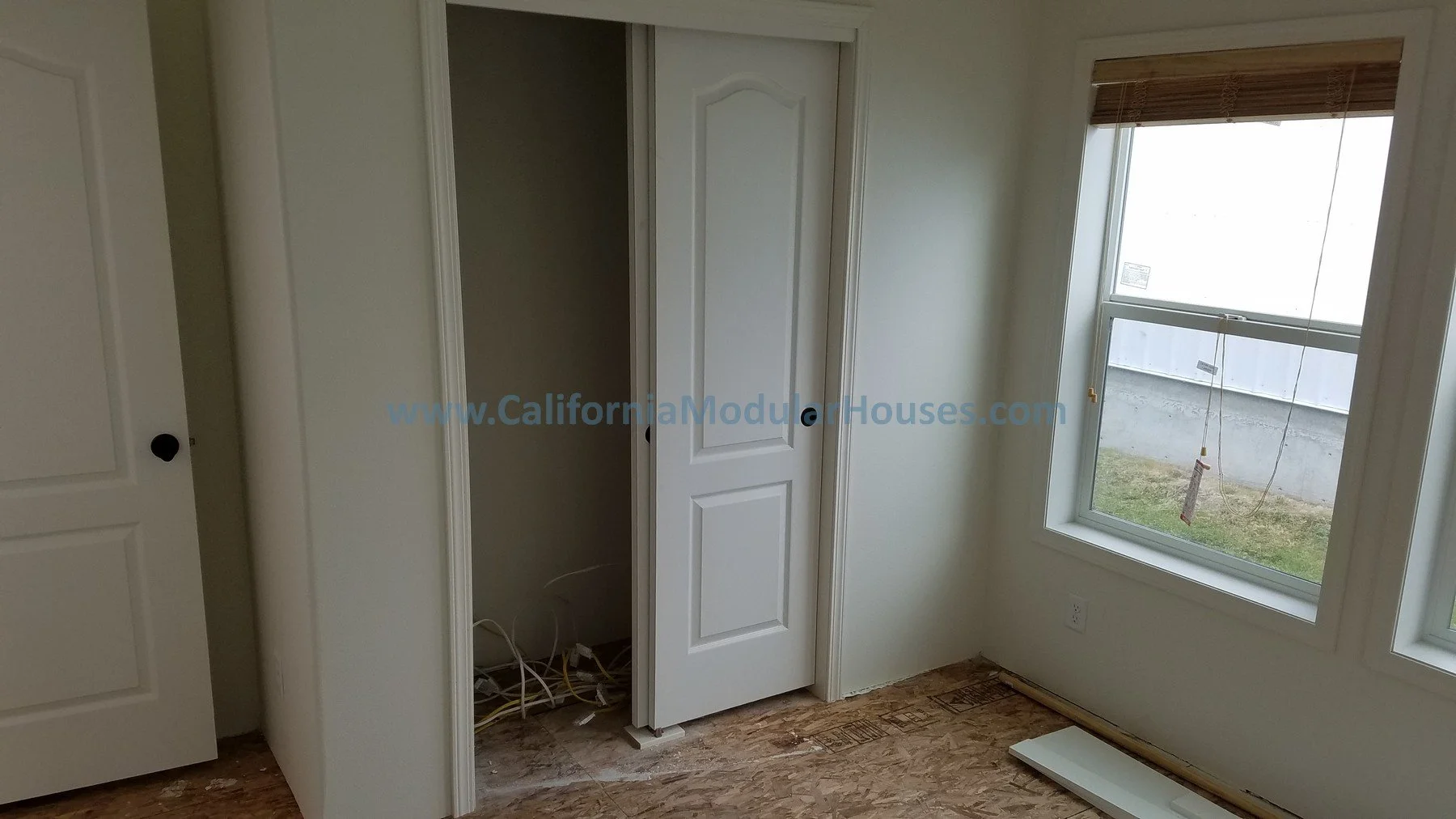 Empty room with white walls, a closet with an open door, a window with blinds, and exposed wiring on the floor. Modular Homes in California, Modular Homes California, Modular Homes, California Modular Home.  