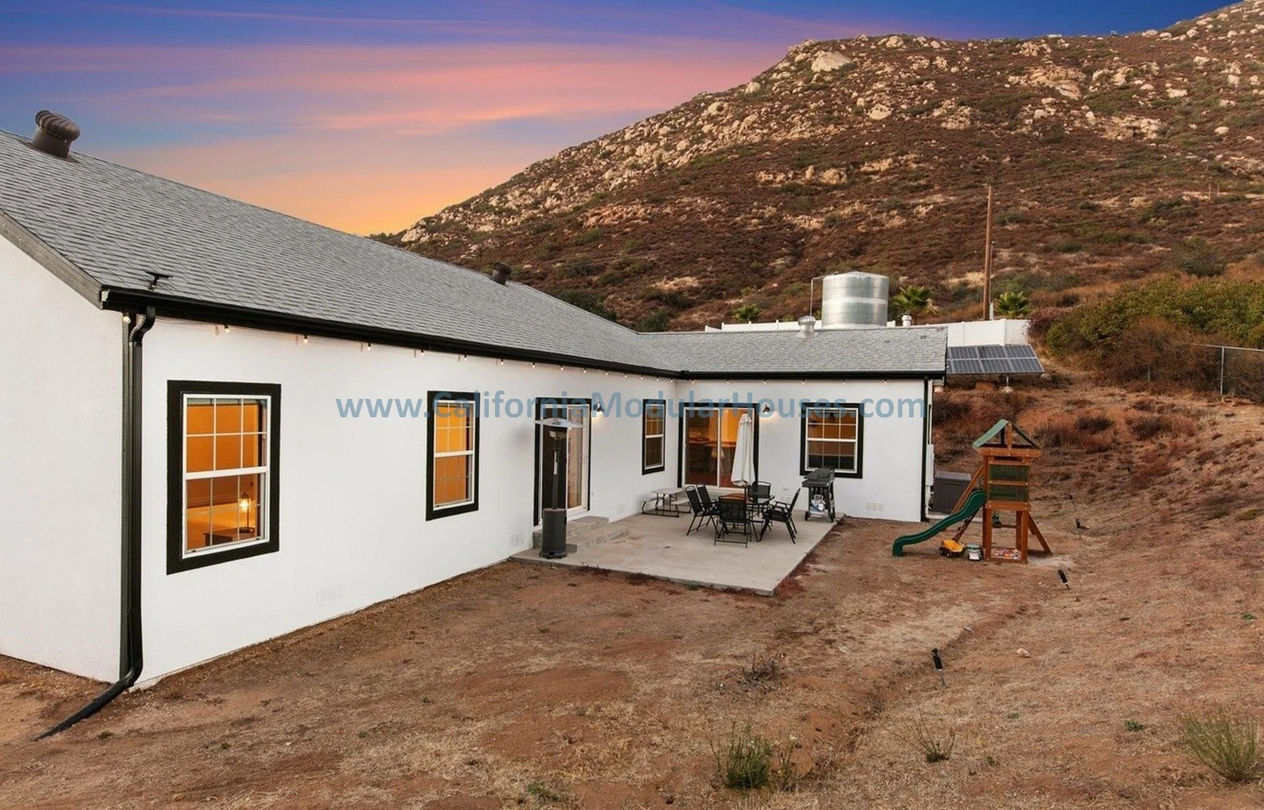A modern white house with black window frames is set against a hilly landscape at sunset. The backyard features a patio with outdoor furniture, a barbecue grill, a children's playset, and solar panels on the roof.