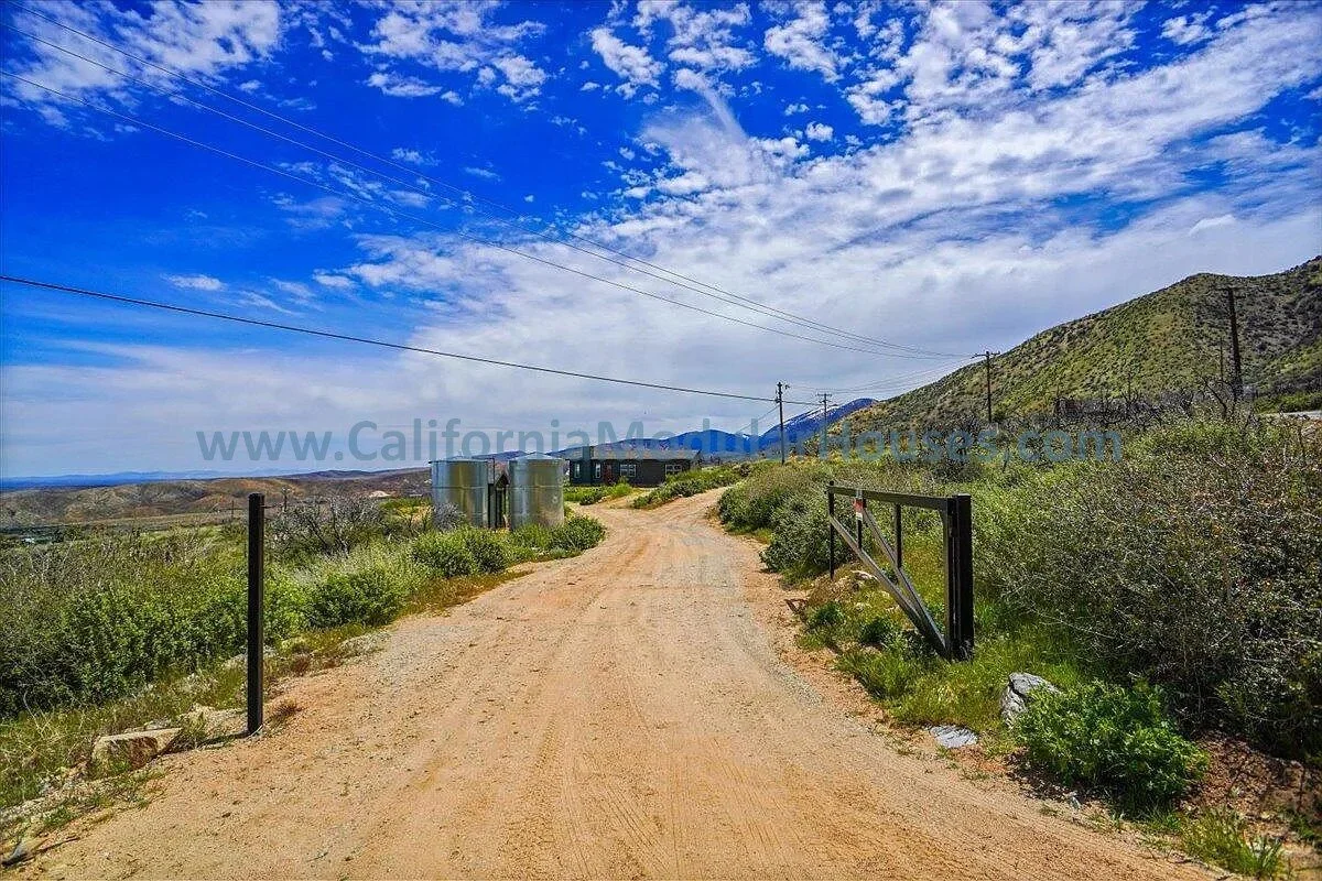 A dirt road winding through a hilly landscape with green shrubs and grass, leading towards a rural house in the distance under a bright blue sky with scattered white clouds.