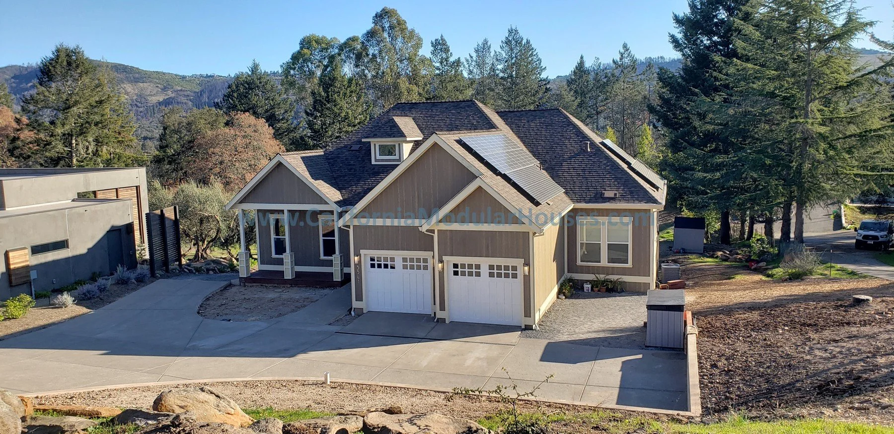 A newly constructed house with solar panels on the roof, a concrete driveway, and a surrounding landscape of trees and mountains. Modular Home CA, Prefab Modular California, California Modular Home,