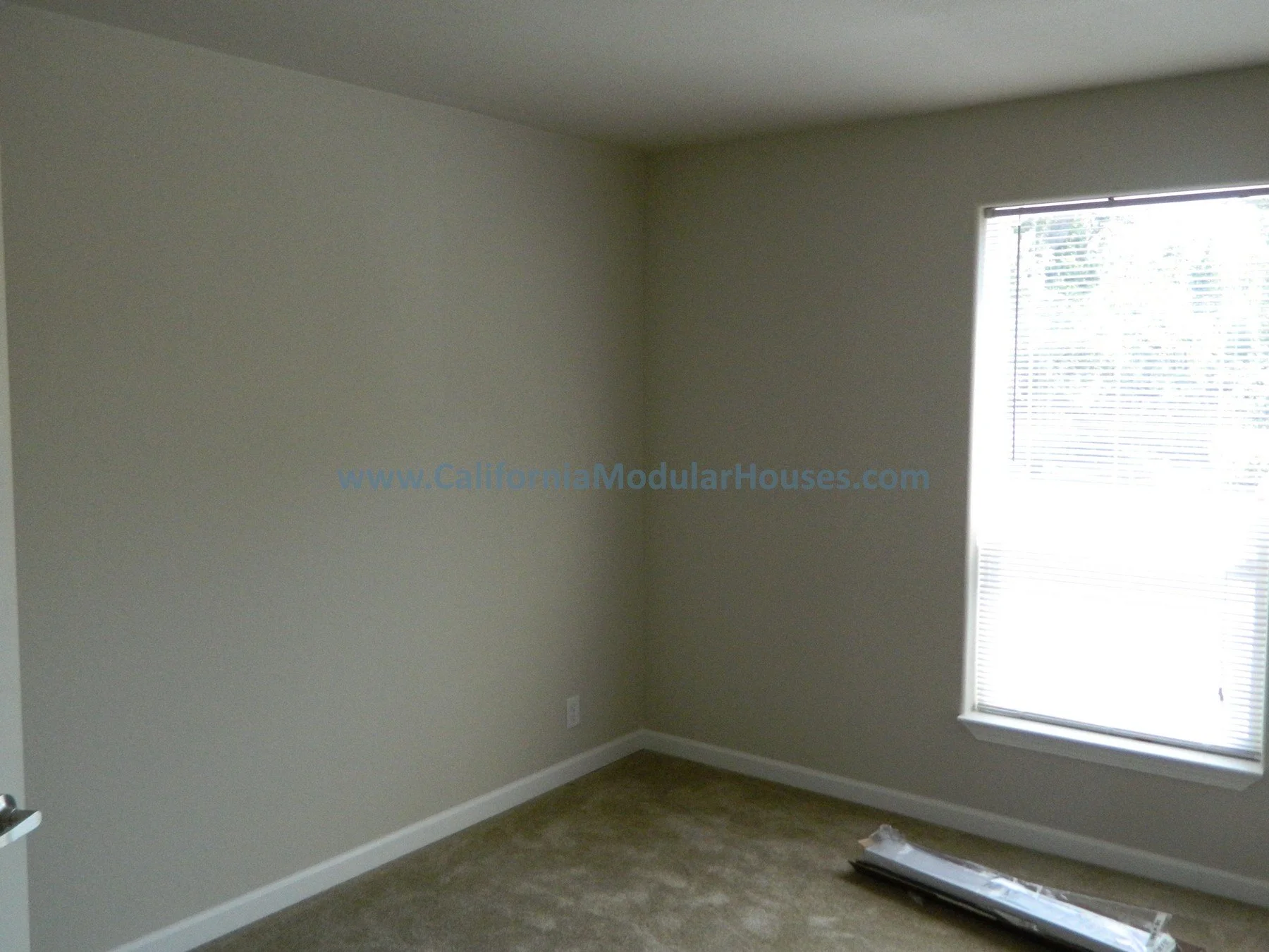 Empty room with beige walls, a window with blinds, beige carpet, and a boxed item on the floor.