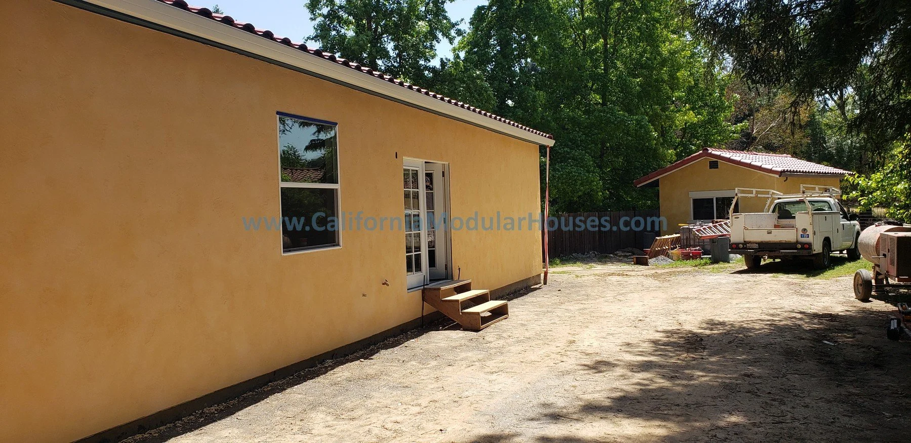 Backyard view of a yellow stucco ADU with a white door and two windows, surrounded by trees, with a smaller yellow building in the background.  Accessory Dwelling Unit for Healdsburg, CA.  Sonoma County Modular Backyard ADU.  