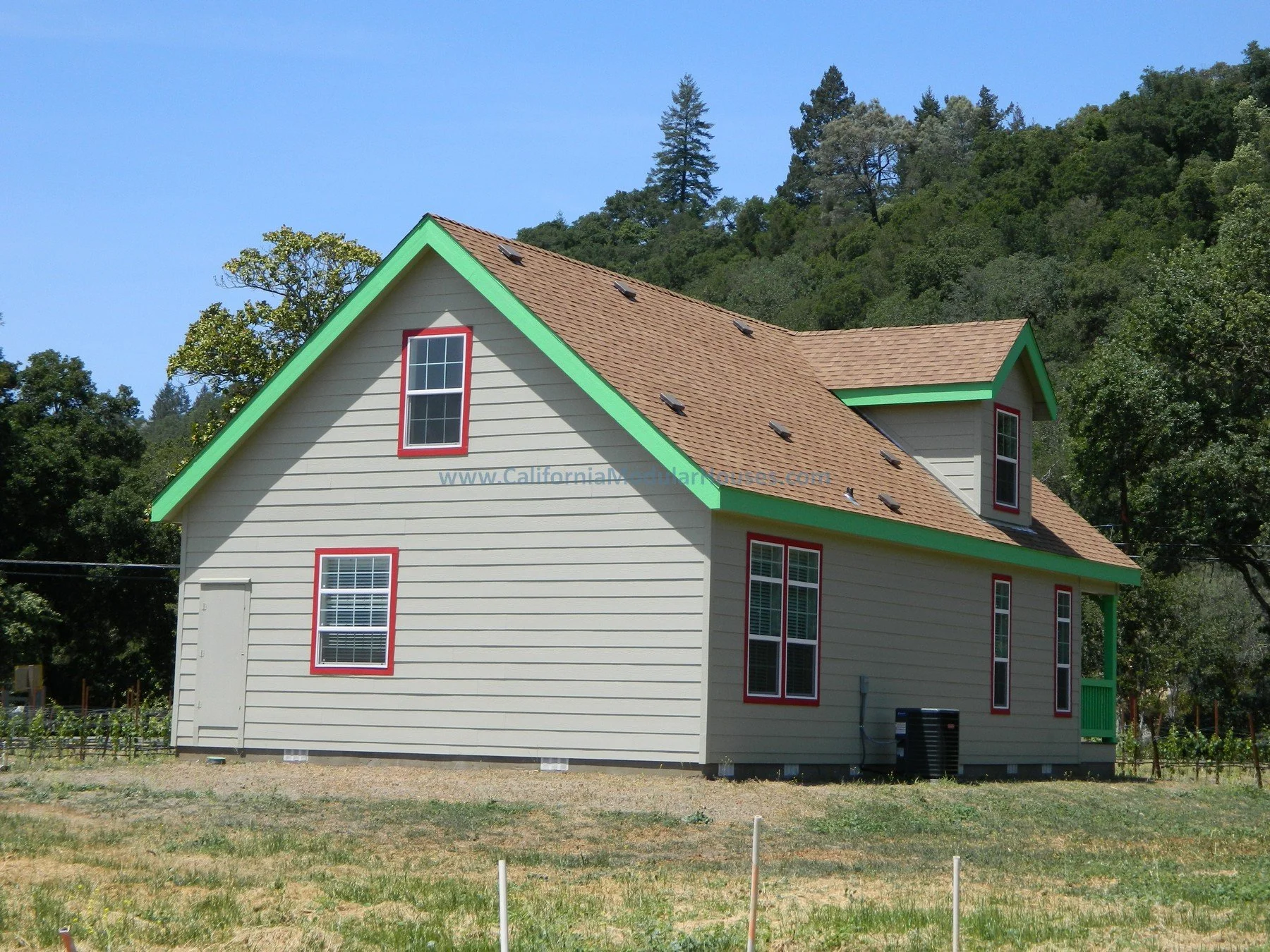 A two-story house with cream-colored siding and red window frames, green trim on the roof edges, and a small front porch, situated in a grassy yard with trees and a hillside in the background under a clear blue sky.
