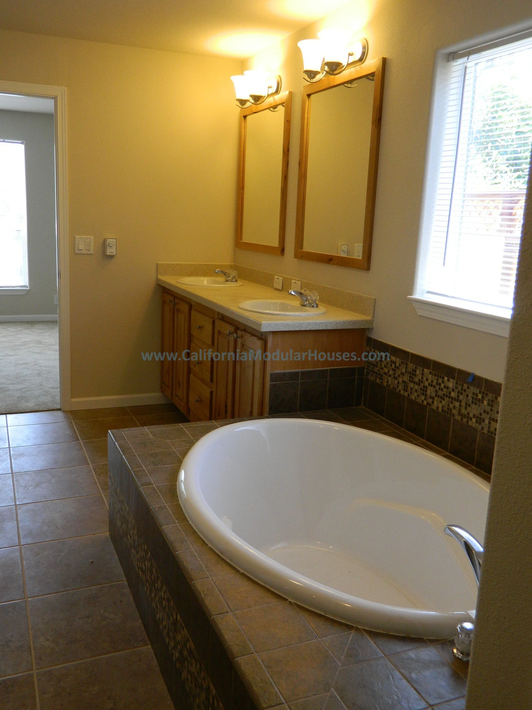 Bathroom with a large bathtub, double sink vanity with wooden cabinets and mirrors, and a window with blinds.