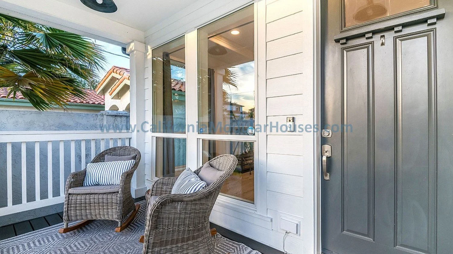 Balcony entrance with two wicker chairs with pillows, white railing, glass sliding door, gray front door, and palm trees in the background.  Pre-Fab brand new home.  custom design.  Tall windows.  Craftsman front door.  