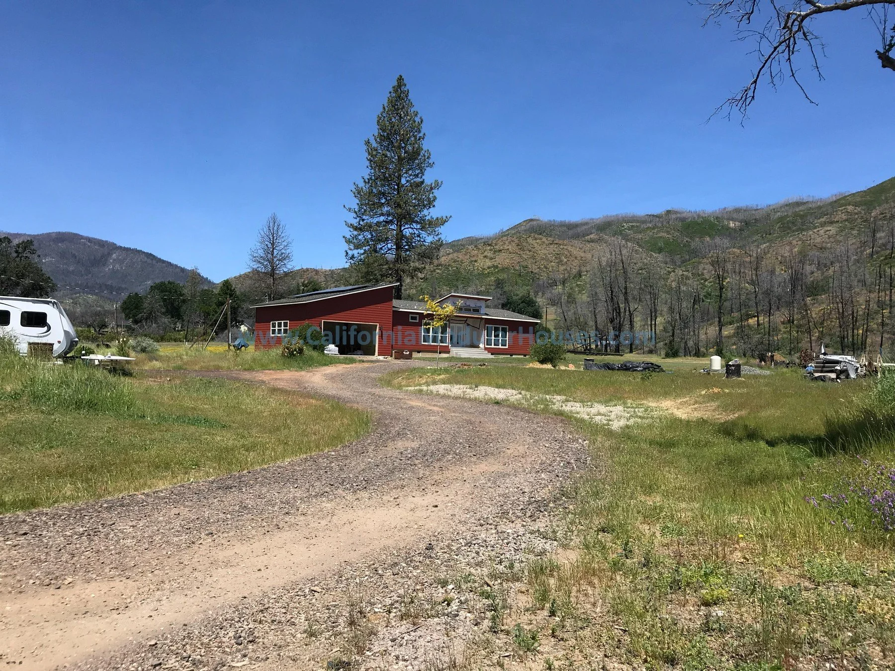 A red modern house with large windows sits in a grassy area with a gravel driveway leading up to it, surrounded by trees and hills under a clear blue sky. Modular Home CA, California Modular Homes.