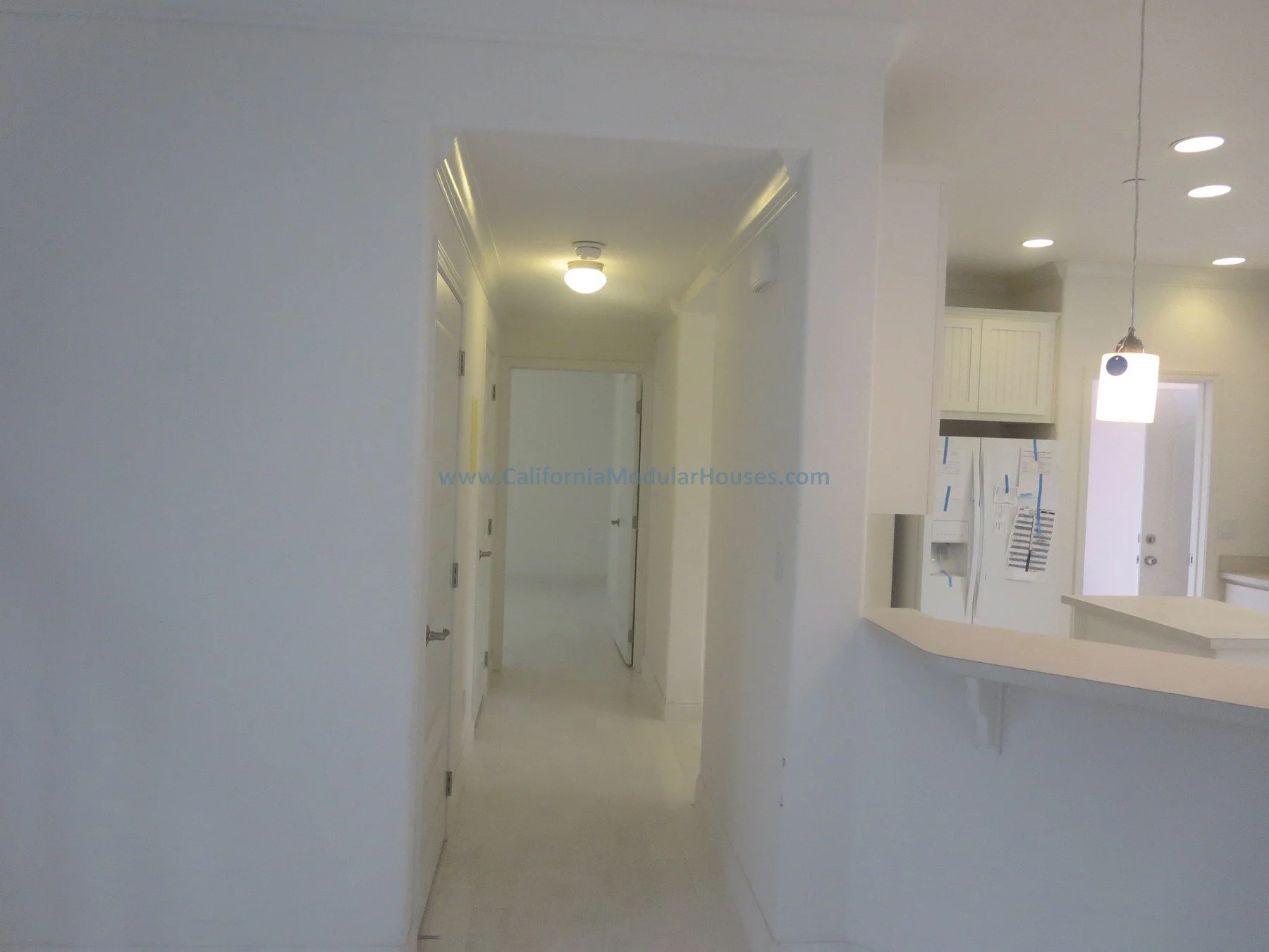 Interior view of a modern, white kitchen with a breakfast bar, hanging pendant light, and ceiling spotlights, seen through an open wall to a hallway with closed doors.  Unfinished attic.  New prefab modular house in Napa, CA.  California modular home