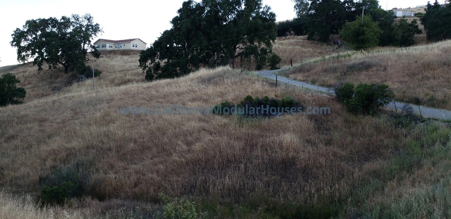 A hilly landscape with dry grass, trees, and a winding dirt road leading up a hill. A house is visible at the top of the hill, and there is a www.CaliforniaModularHouses.com watermark across the image. California Modular,